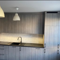 Włodarczyk Meble - Sandra Włodarczyk - Nowoczesna kuchnia z szarymi frontami, czarnym blatem i minimalistycznymi uchwytami. Oświetlenie podszafkowe podkreśla dekoracyjne płytki. Dwie lampy wiszące nad zlewem.