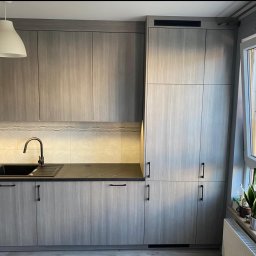 Włodarczyk Meble - Sandra Włodarczyk - Nowoczesna kuchnia na wymiar z szarymi, drewnopodobnymi frontami, czarnym zlewem i blatem. Minimalistyczny design, oświetlenie LED pod szafkami, roślina w oknie.