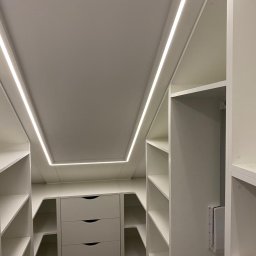Włodarczyk Meble - Sandra Włodarczyk - Jasna garderoba na poddaszu z oświetleniem LED. Widok z dołu ukazuje półki i szuflady. Minimalistyczny design, białe wykończenie, nowoczesne oświetlenie.