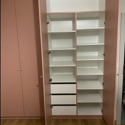 Włodarczyk Meble - Sandra Włodarczyk - Różowa szafa z otwartymi drzwiami, ukazująca białe półki i szuflady. Widoczne okucia i drewniana podłoga. Minimalistyczny design, jasne wnętrze szafy.