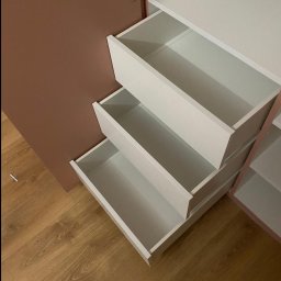 Włodarczyk Meble - Sandra Włodarczyk - Wnętrze szafy z trzema wysuniętymi, białymi szufladami na tle brązowej ściany i drewnianej podłogi. Widoczne zawiasy i półki wewnątrz szafy.