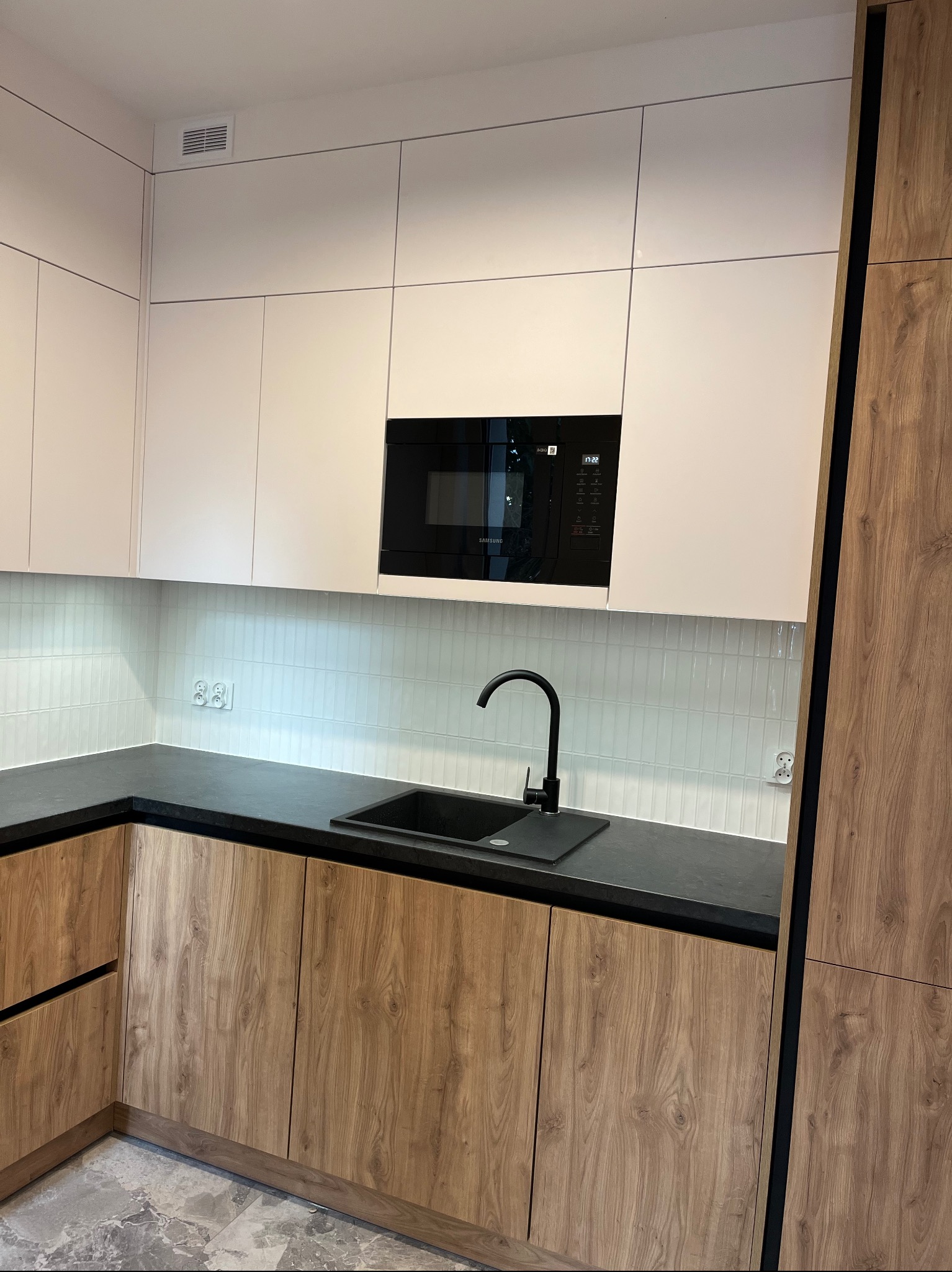 Nowoczesna kuchnia z drewnianymi frontami szafek dolnych i białymi górnymi. Czarny blat i armatura, białe płytki na ścianie. Wbudowana mikrofalówka Samsung.