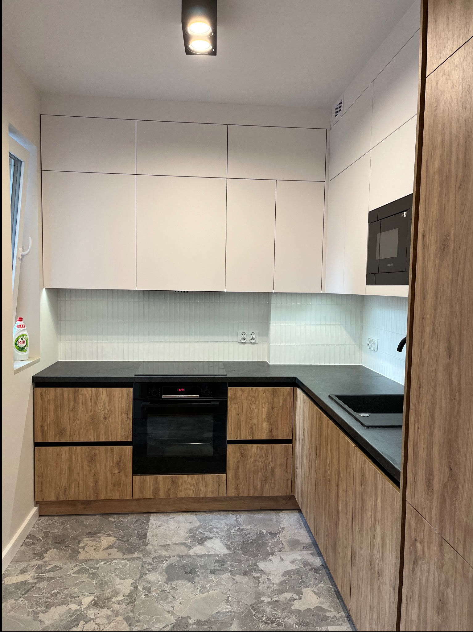 Nowoczesna kuchnia z drewnianymi frontami szafek dolnych i białymi górnymi, czarny blat i sprzęt AGD, płytki na ścianie w kształcie cegiełek i marmurowa podłoga.