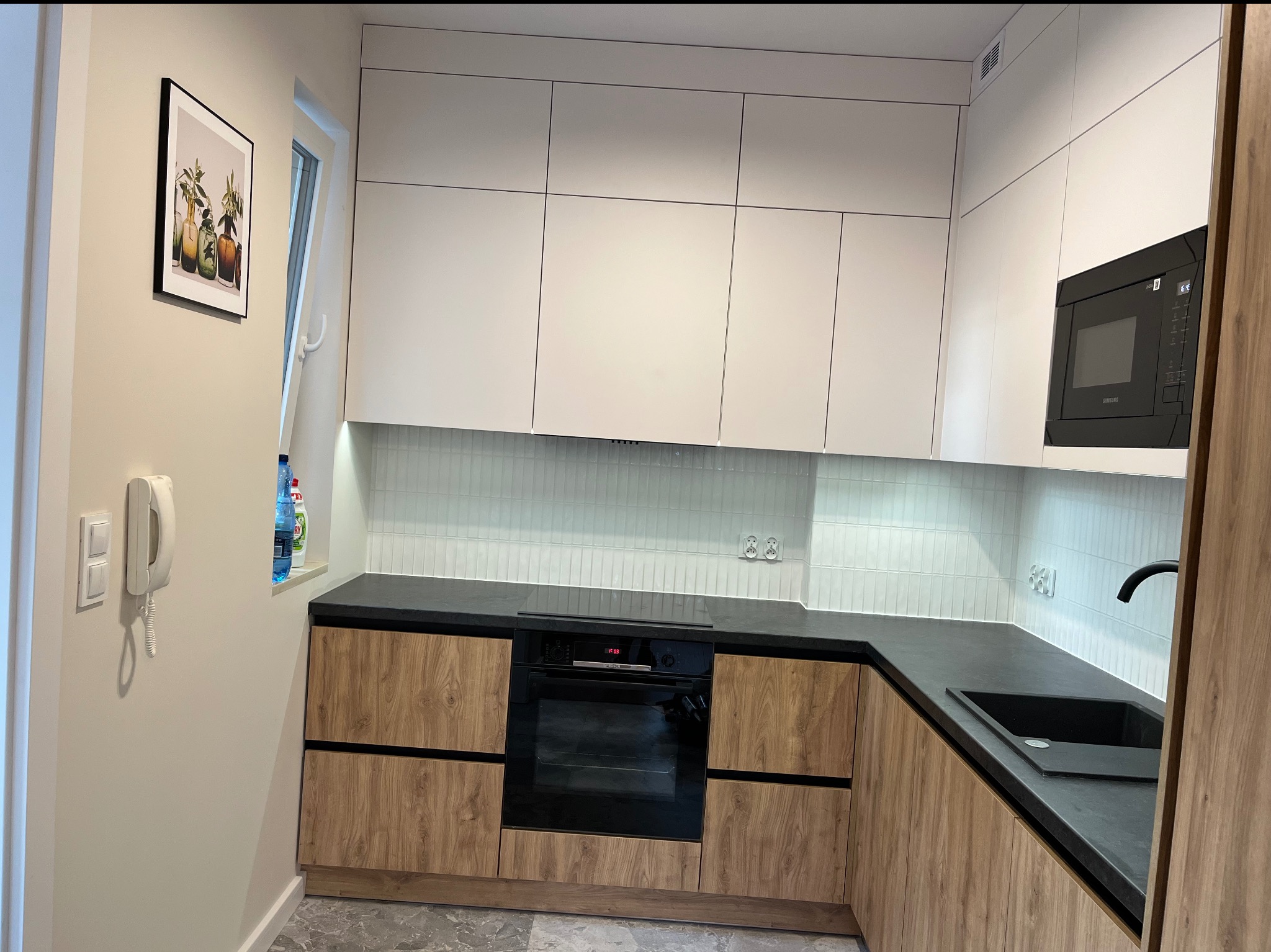 Nowoczesna kuchnia z białymi szafkami górnymi i drewnopodobnymi dolnymi. Czarny blat i subtelne oświetlenie LED podkreślają minimalistyczny design. Uwagę przykuwa obraz na ścianie.