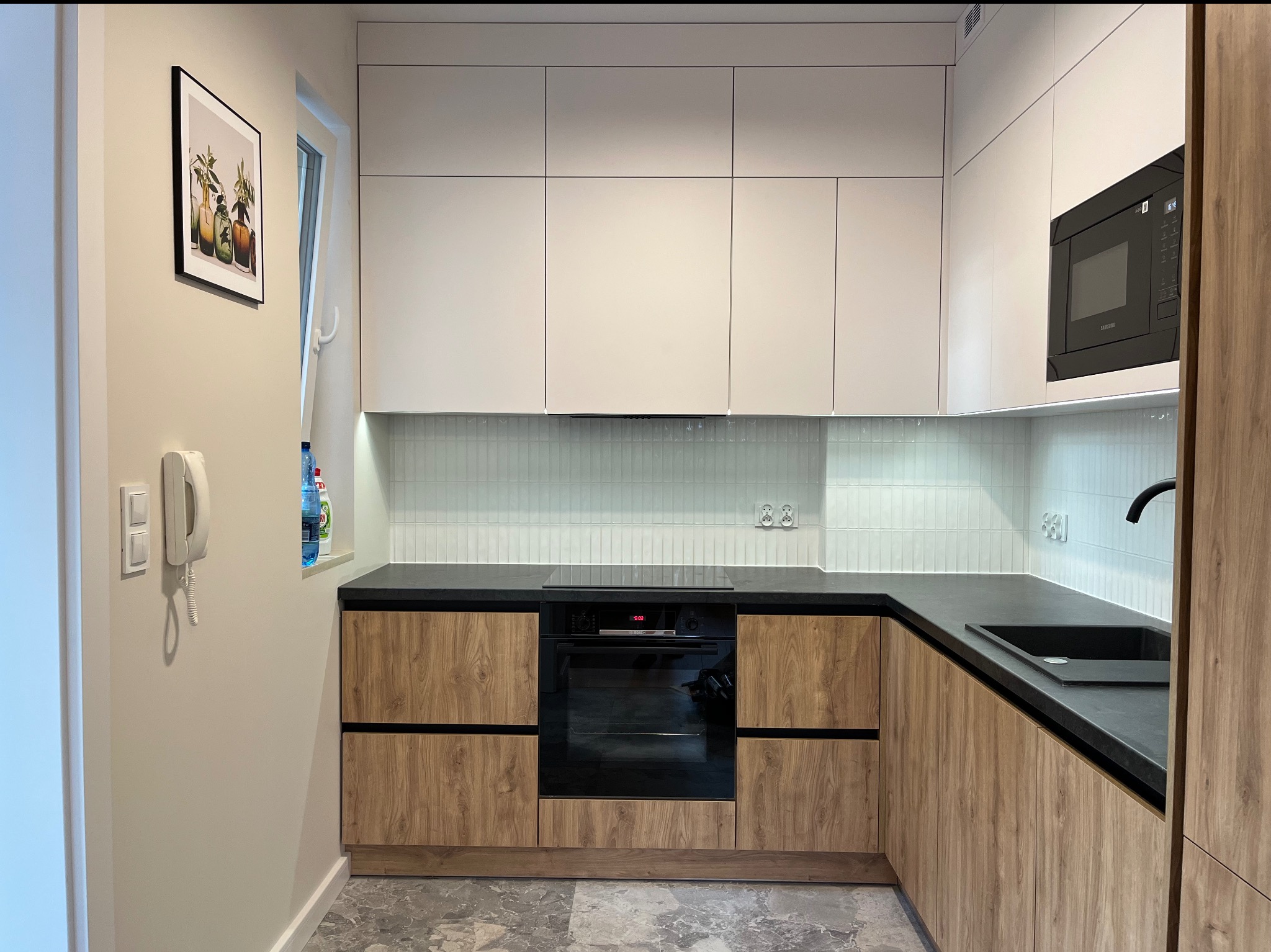 Nowoczesna kuchnia z białymi szafkami górnymi i dolnymi z drewnianym wzorem. Czarny blat, piekarnik i mikrofalówka. Na ścianie wisi obrazek w czarnej ramce.