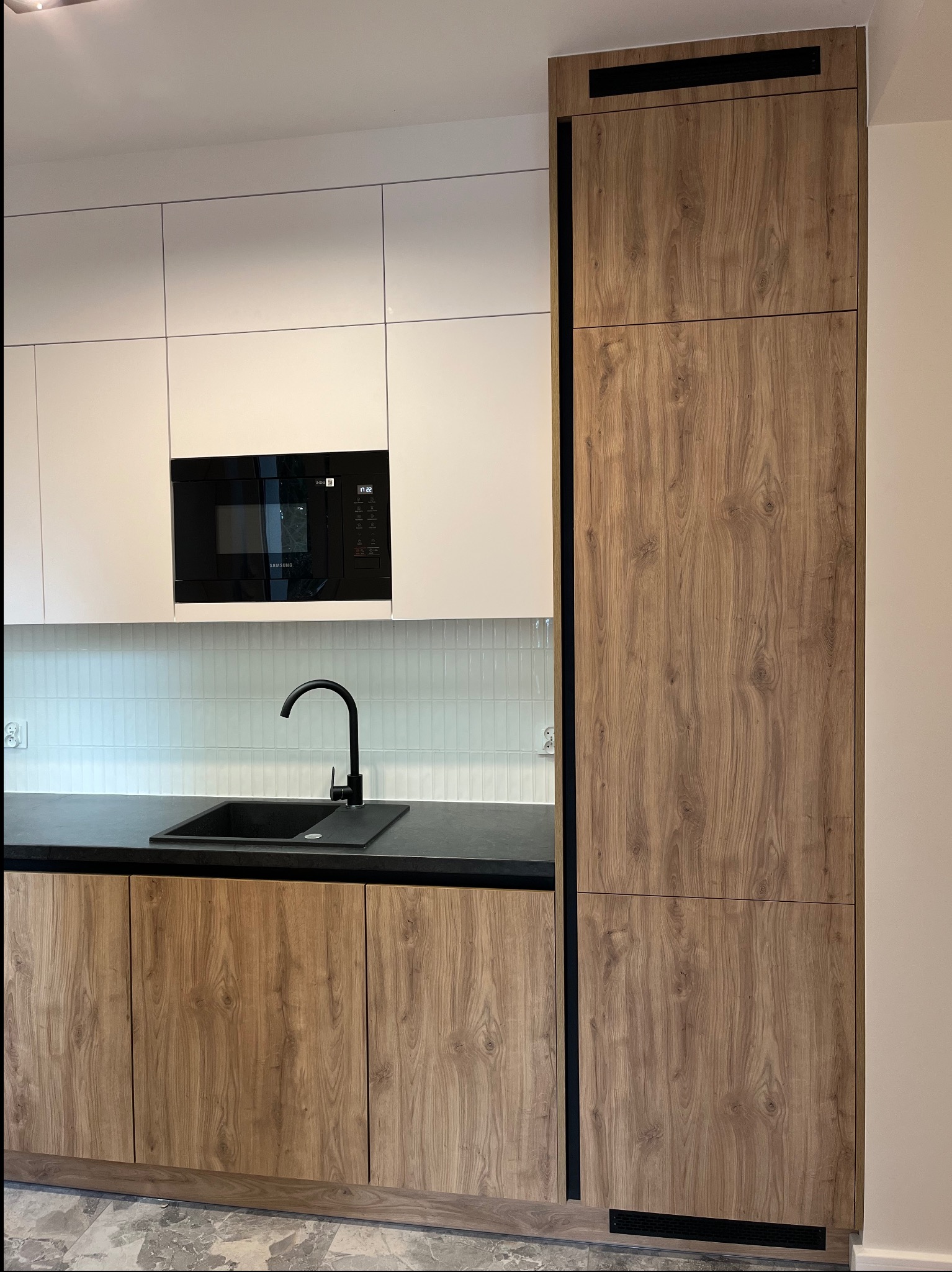 Nowoczesna kuchnia z frontami drewnopodobnymi i białymi szafkami. Czarny zlew i bateria, podświetlane płytki. Wysoka szafa z czarnymi akcentami.