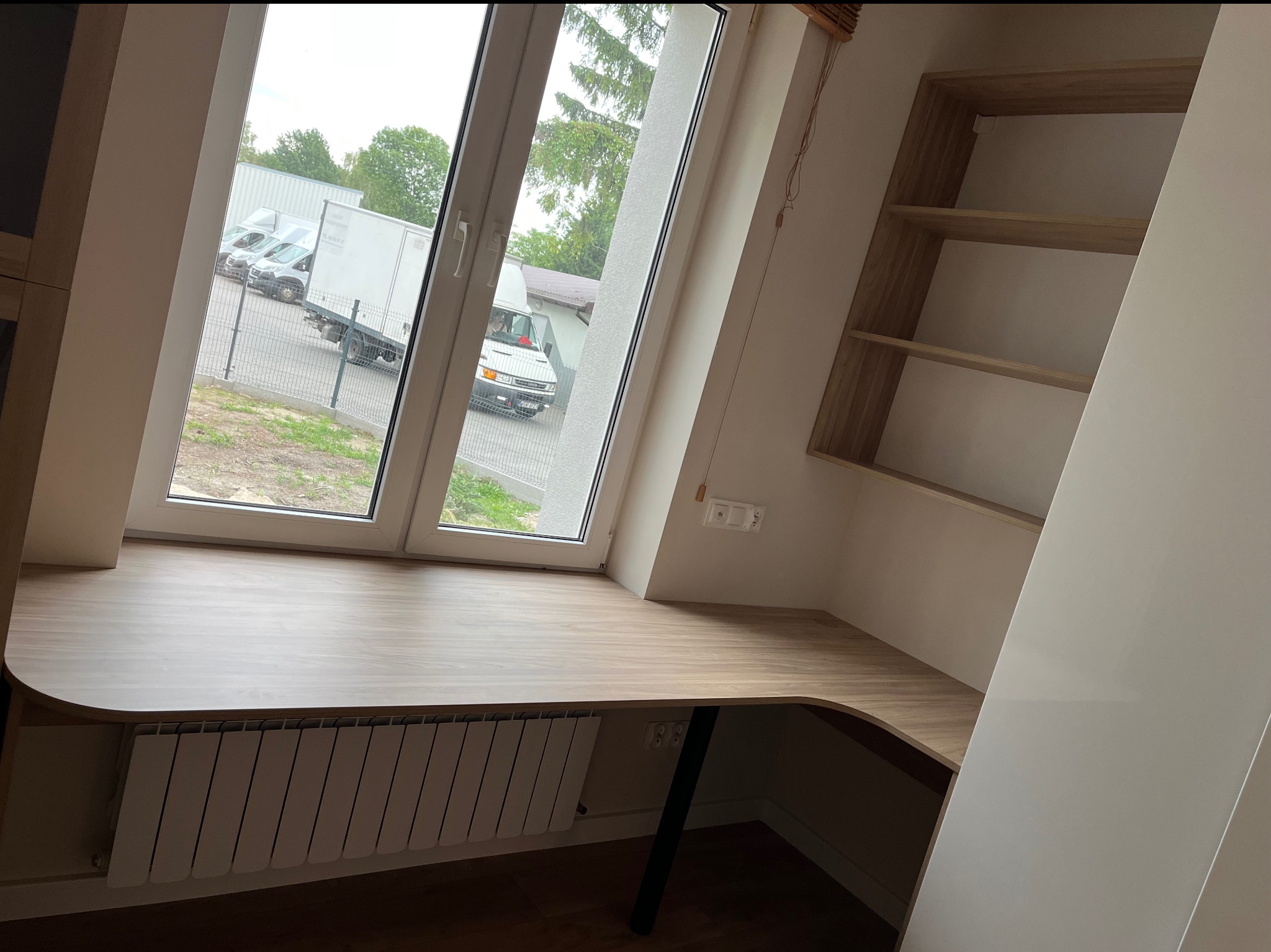Drewniany blat biurka na wymiar w pokoju na poddaszu, zintegrowany z grzejnikiem i półkami wnękowymi. Widok z okna na samochody. Jasne wnętrze.