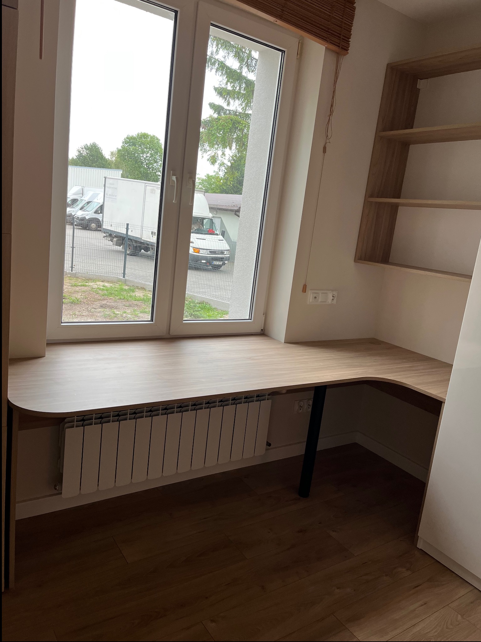 Drewniany blat biurka na wymiar pod oknem, z grzejnikiem pod spodem. Na ścianie półki z tego samego materiału. Widok z okna na samochody dostawcze.