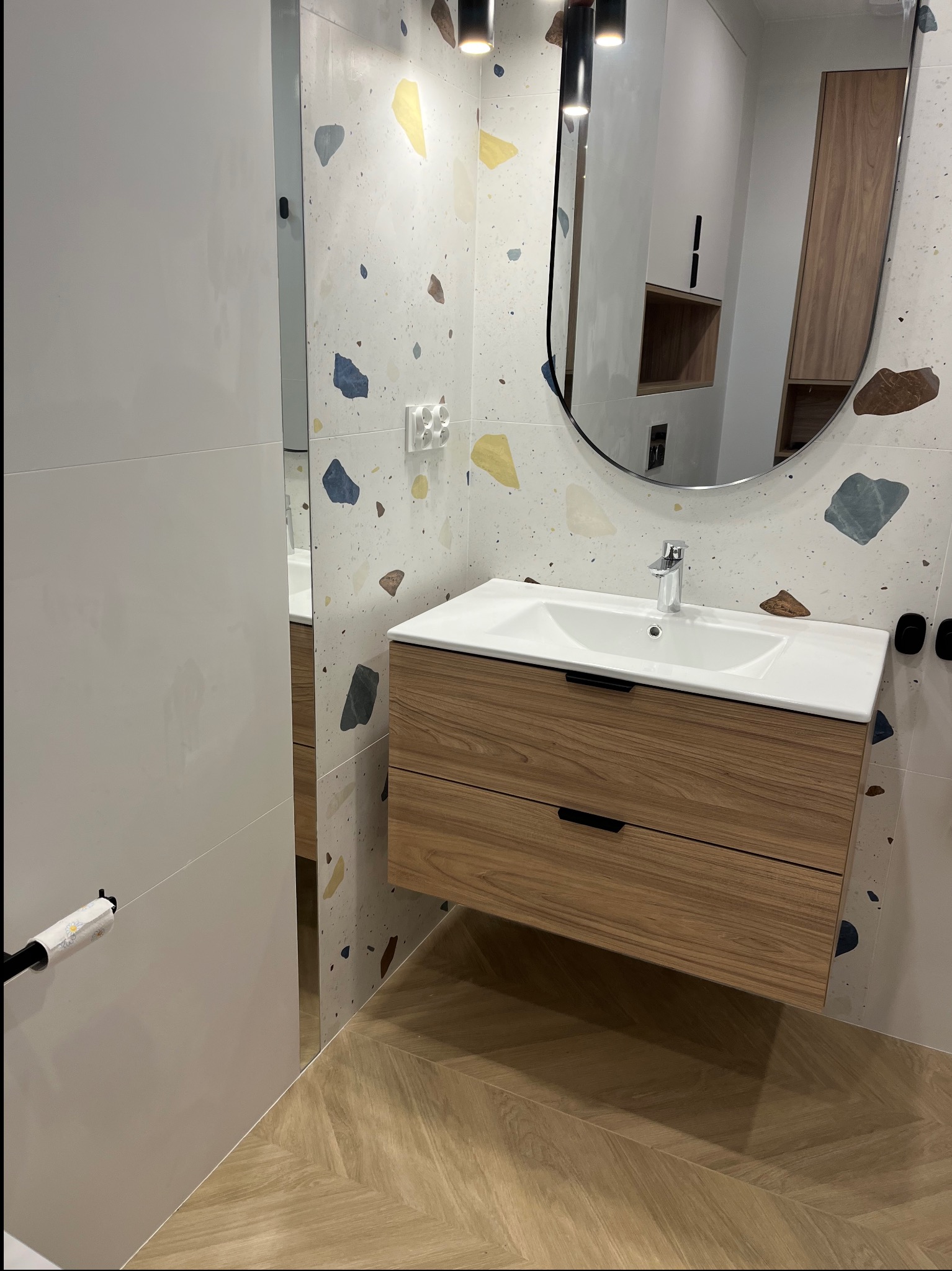 Nowoczesna łazienka z drewnianą szafką pod umywalkę, lustrem w owalnej ramie i płytkami w stylu terrazzo. Widoczny fragment podłogi ułożonej w jodełkę.