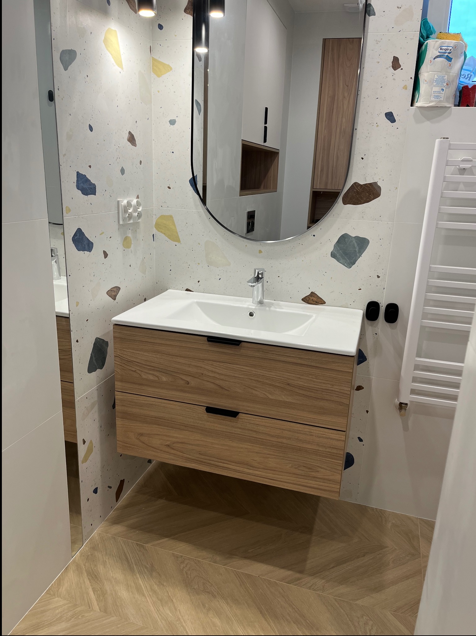 Nowoczesna łazienka z drewnianą szafką pod umywalką, lustrem w owalnej ramie i dekoracyjnymi płytkami w stylu terrazzo na ścianie.