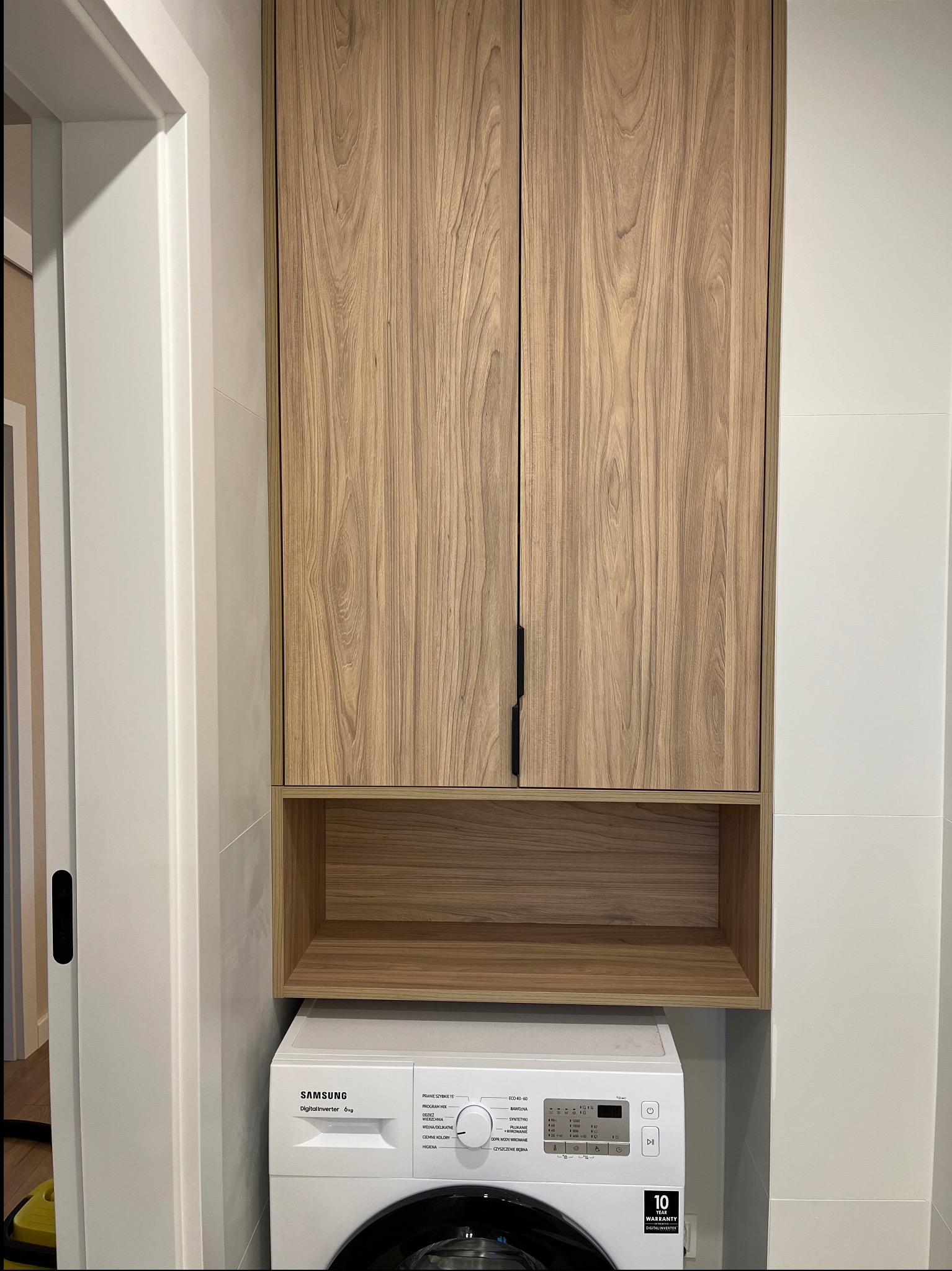 Szafka w łazience z drewnianymi frontami, zamontowana nad pralką. Minimalistyczny design, czarne uchwyty, białe płytki w tle. Praktyczne rozwiązanie do małej łazienki.