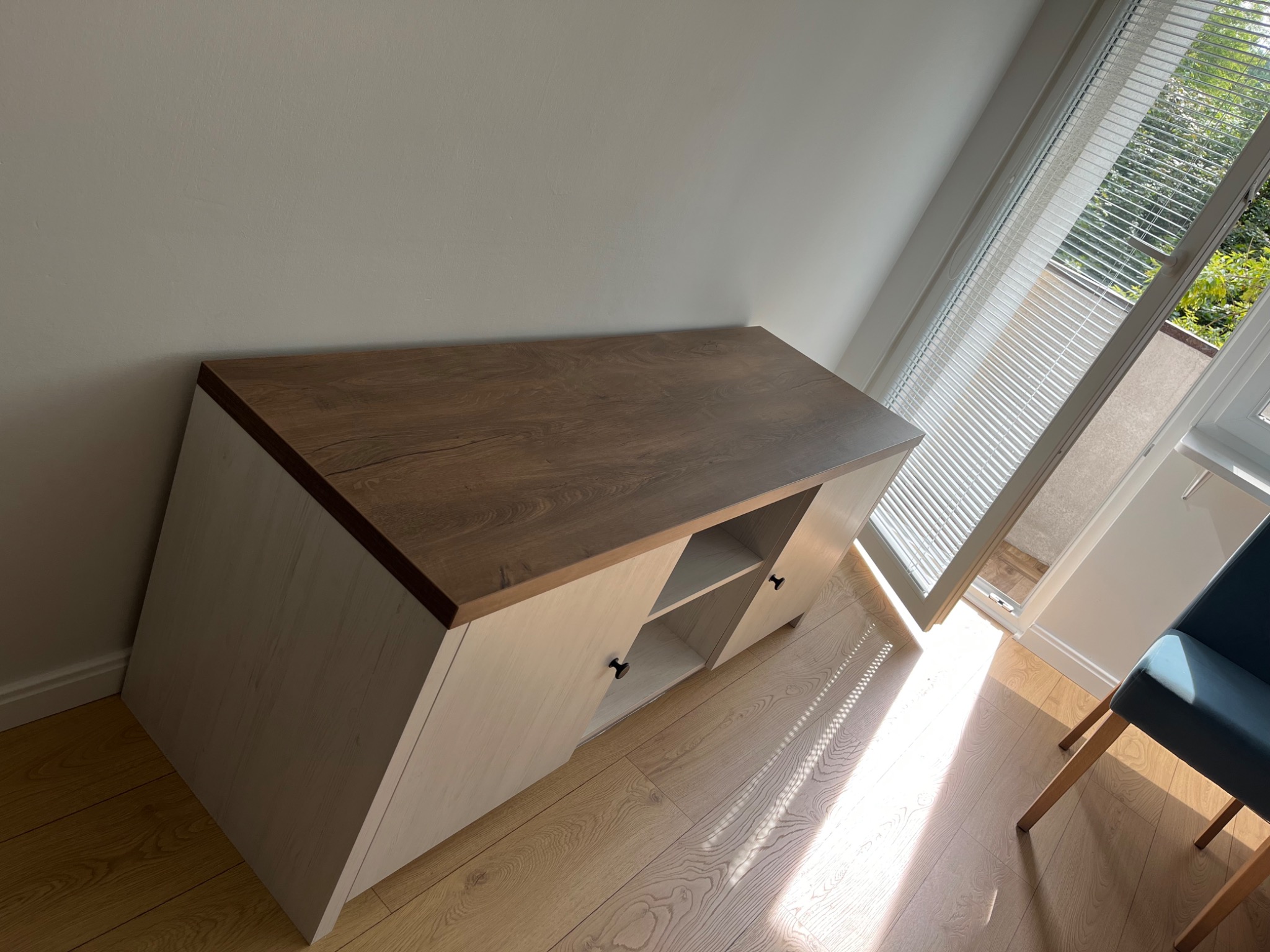 Nowoczesna szafka RTV z drewnianym blatem w jasnym wnętrzu. Minimalistyczny design, widoczne półki i zamknięte szafki. Ustawiona na drewnianej podłodze, blisko okna z żaluzjami.