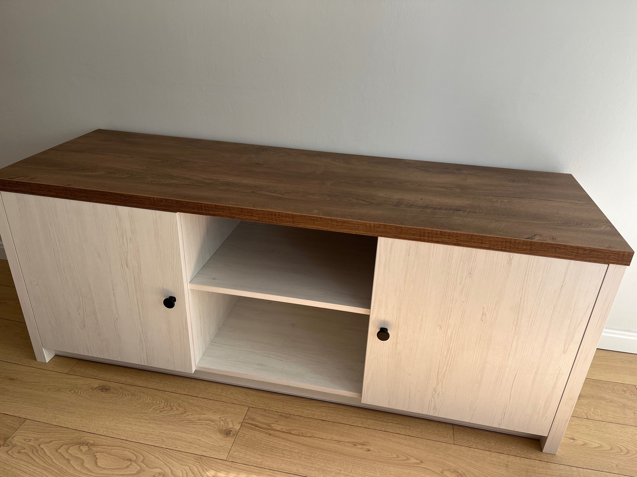 Stojąca szafka RTV z białymi drzwiczkami i blatem w kolorze ciemnego drewna. Widoczne dwie półki w środku. Ustawiona na podłodze z paneli.