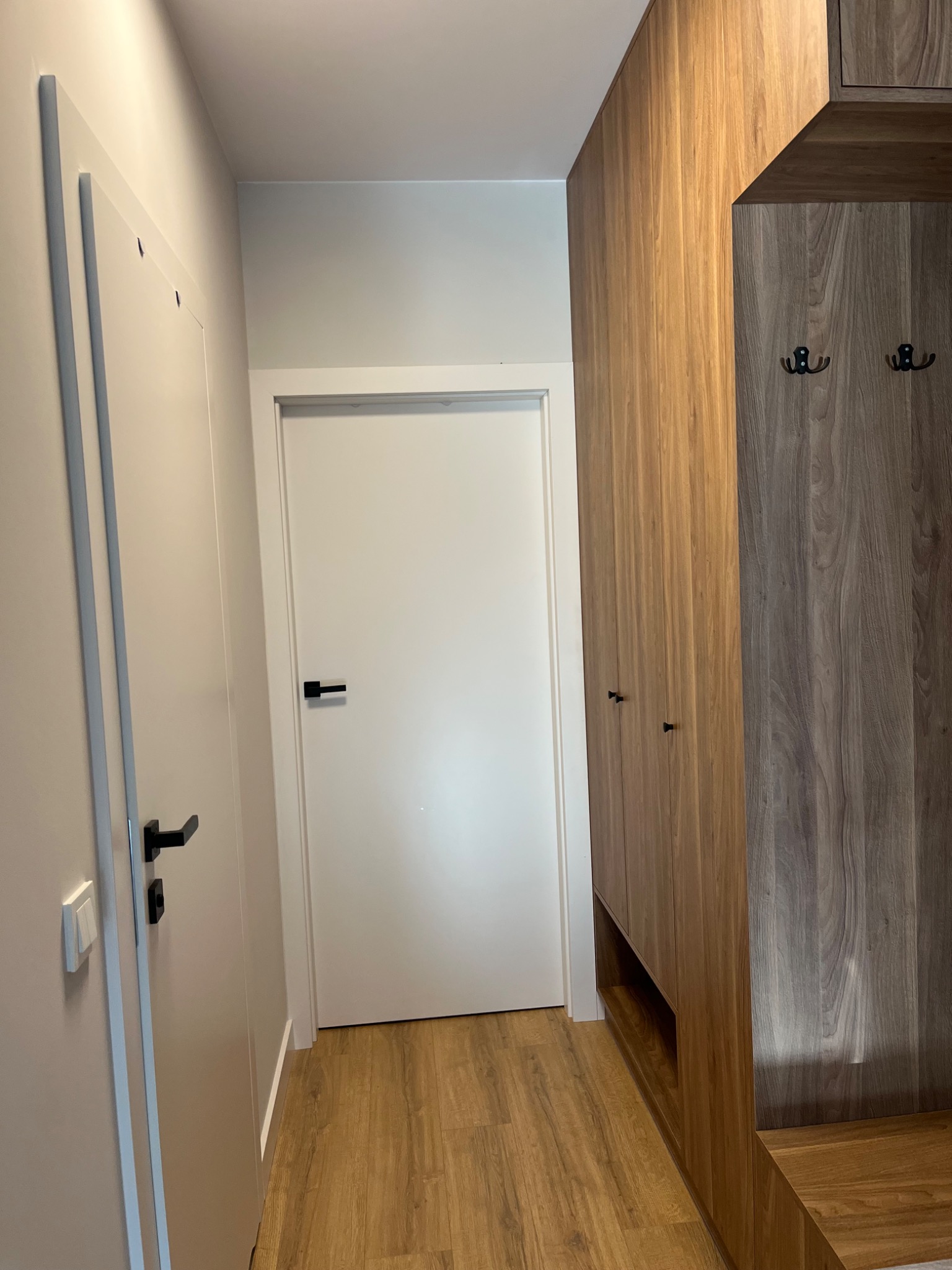 Wnętrze przedpokoju z zabudową szafy w odcieniach drewna i białymi drzwiami. Widoczna podłoga z paneli imitujących drewno i minimalistyczne czarne klamki.