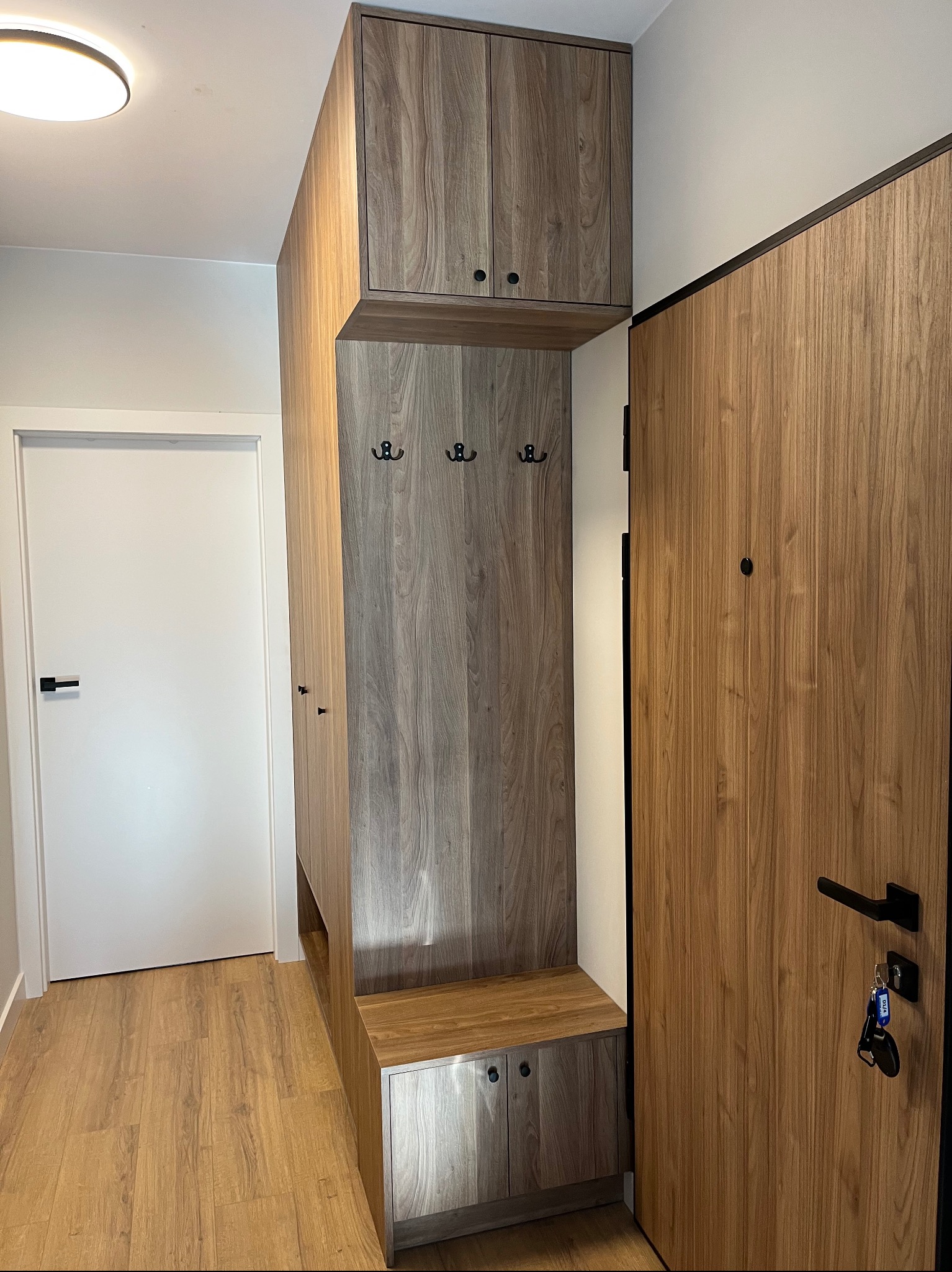 Funkcjonalna zabudowa przedpokoju z drewnopodobnymi frontami, obejmująca szafę, siedzisko i wieszaki. Obok drzwi wejściowe z czarną klamką i kluczami.