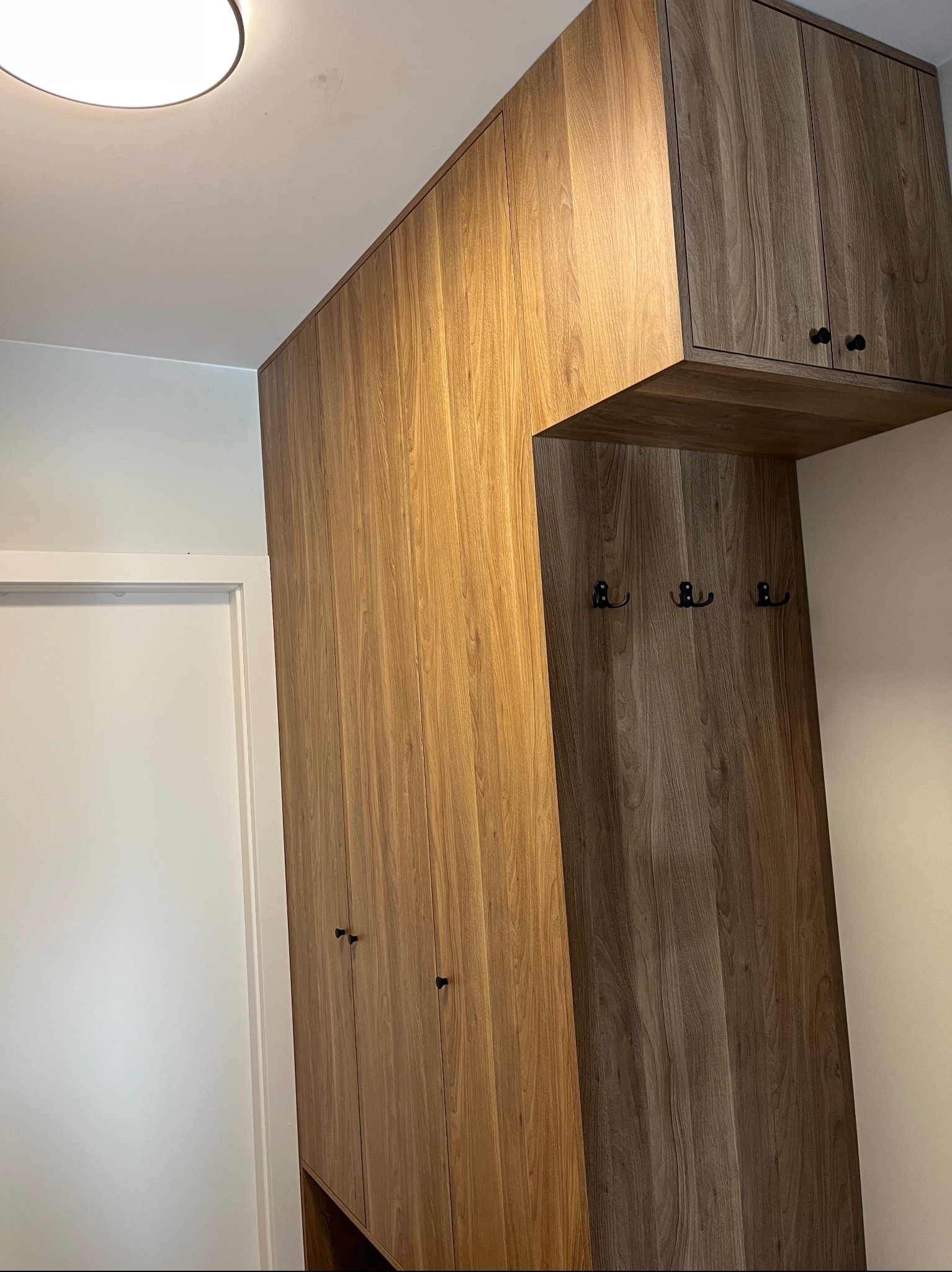 Wysoka szafa wnękowa z drewnianymi frontami i wieszakami, wkomponowana w przedpokój. Minimalistyczne uchwyty, ciepłe oświetlenie sufitowe.