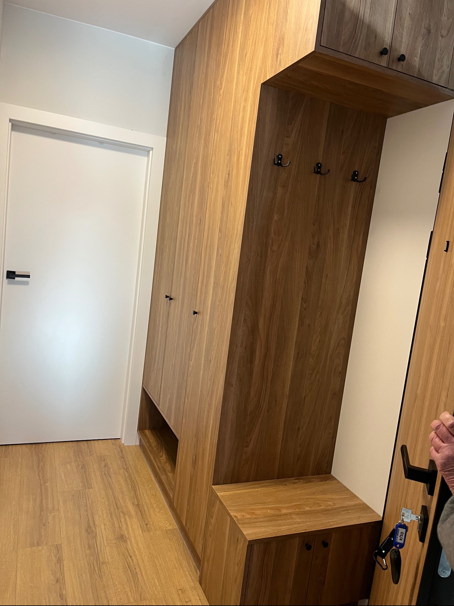 Drewniana szafa wnękowa z siedziskiem i wieszakami w przedpokoju, widok z perspektywy podłogi, drzwi wejściowe po lewej stronie, klucze w zamku.