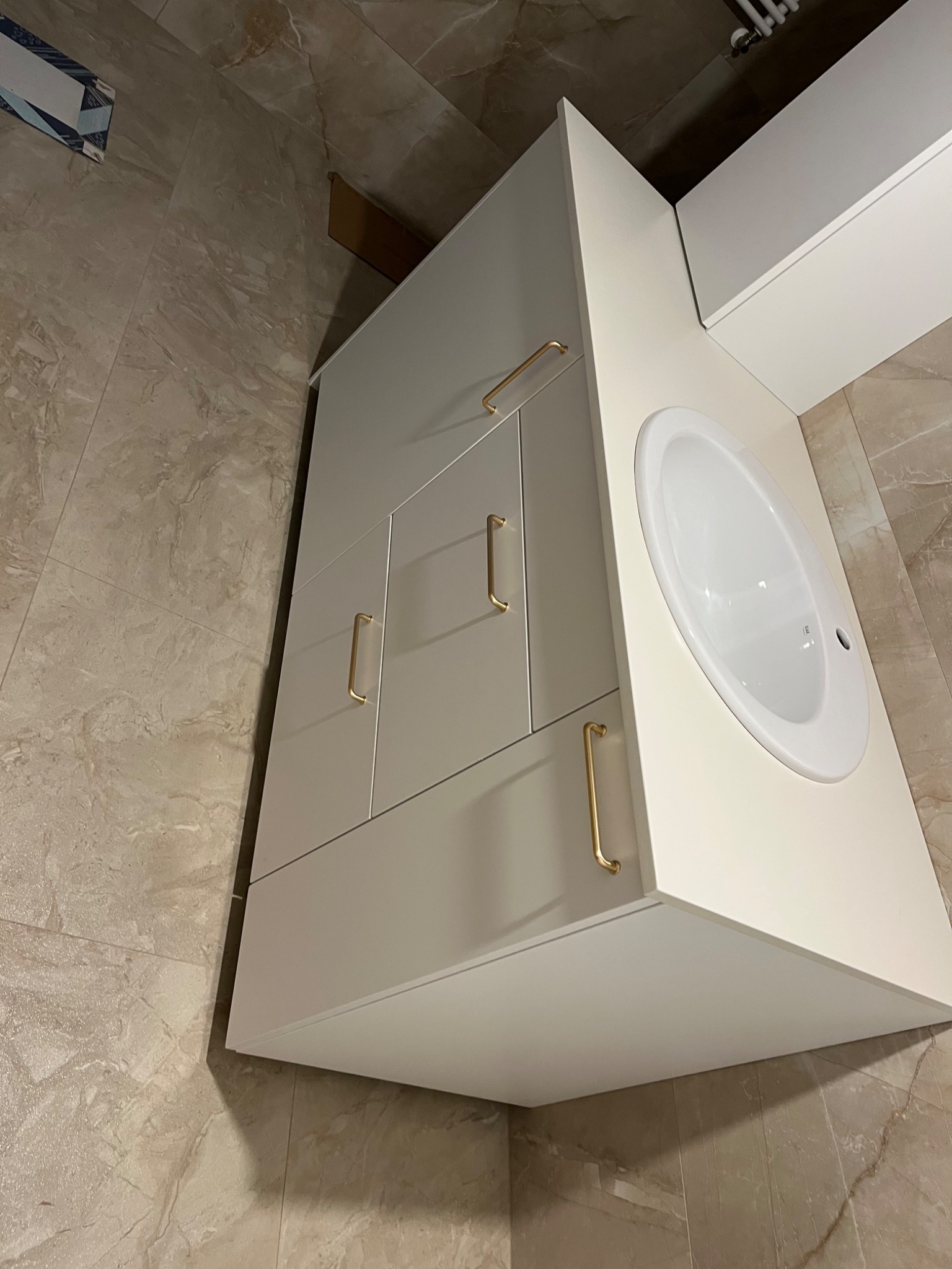 Biała szafka łazienkowa z umywalką i złotymi uchwytami na tle beżowych płytek. Minimalistyczny design, widok z góry, ciepłe oświetlenie.