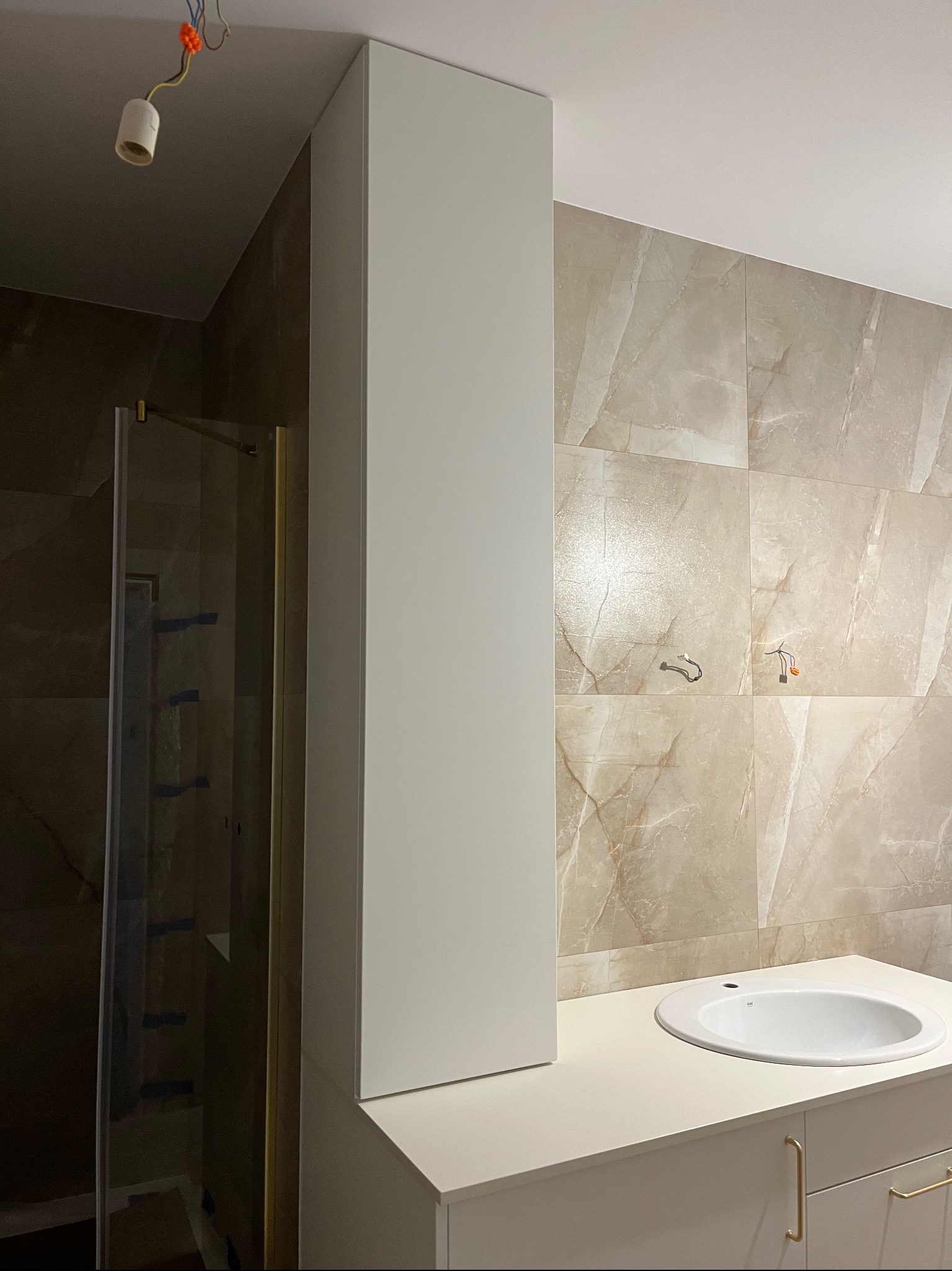 Nowoczesna łazienka z umywalką nablatową i wysoką, białą szafką. Beżowe płytki imitujące marmur na ścianie. Widoczna kabina prysznicowa z złotymi profilami.