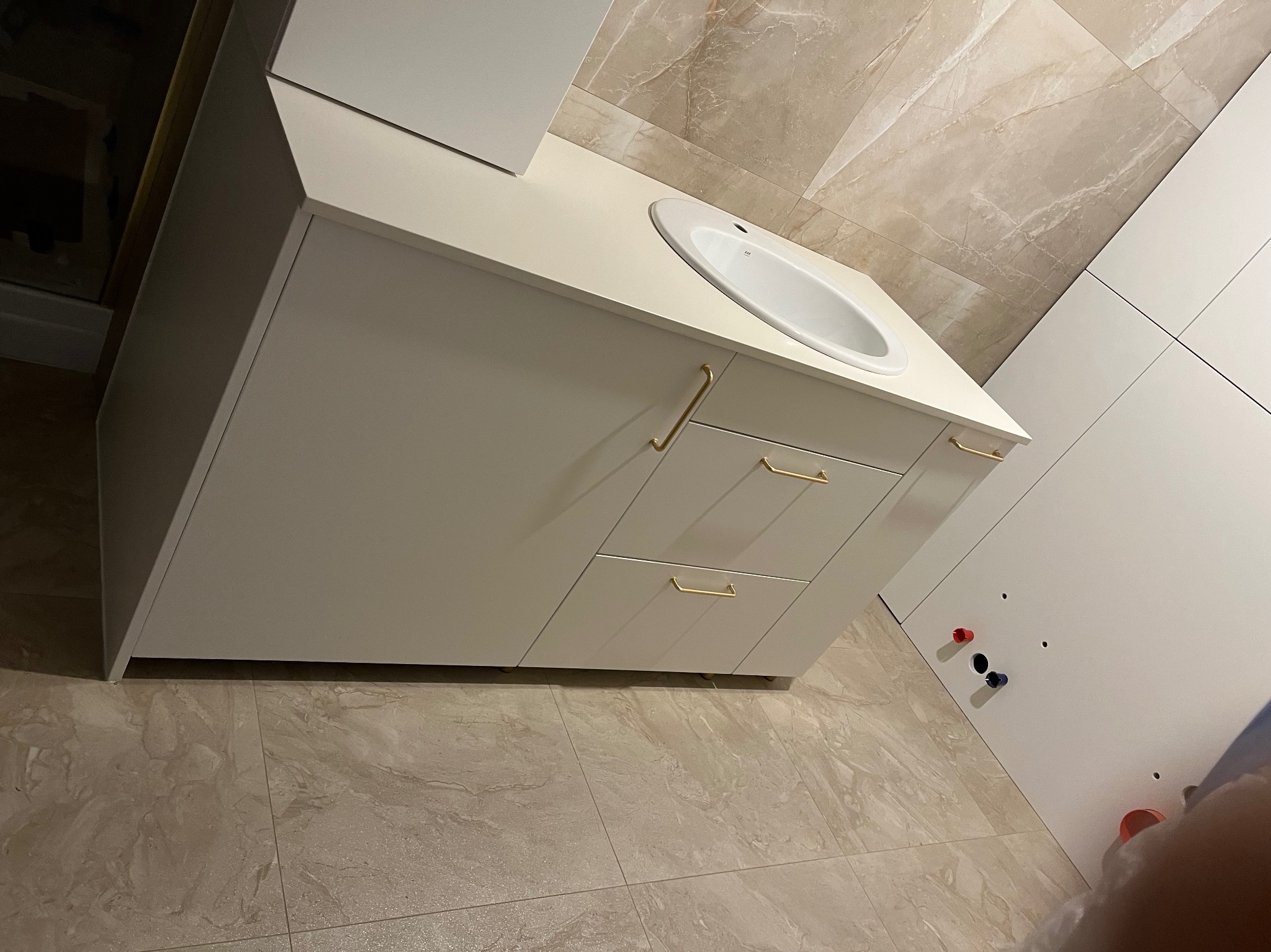 Biała szafka łazienkowa z umywalką i złotymi uchwytami na tle beżowych płytek imitujących marmur. Widoczne przyłącza wodne.