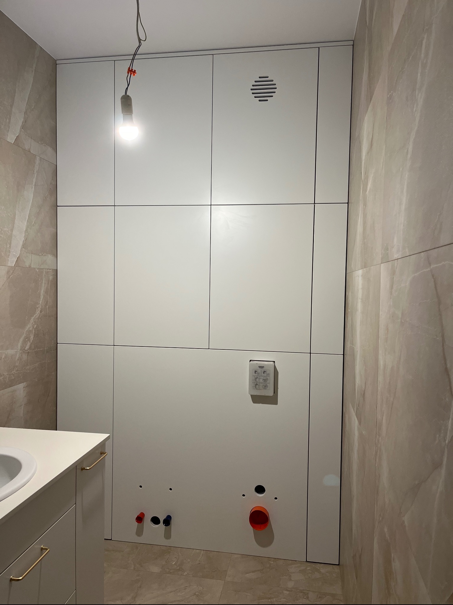 Biała zabudowa w łazience z widocznymi przyłączami hydraulicznymi i panelem sterowania, oświetlona żarówką na kablu. Minimalistyczny design.