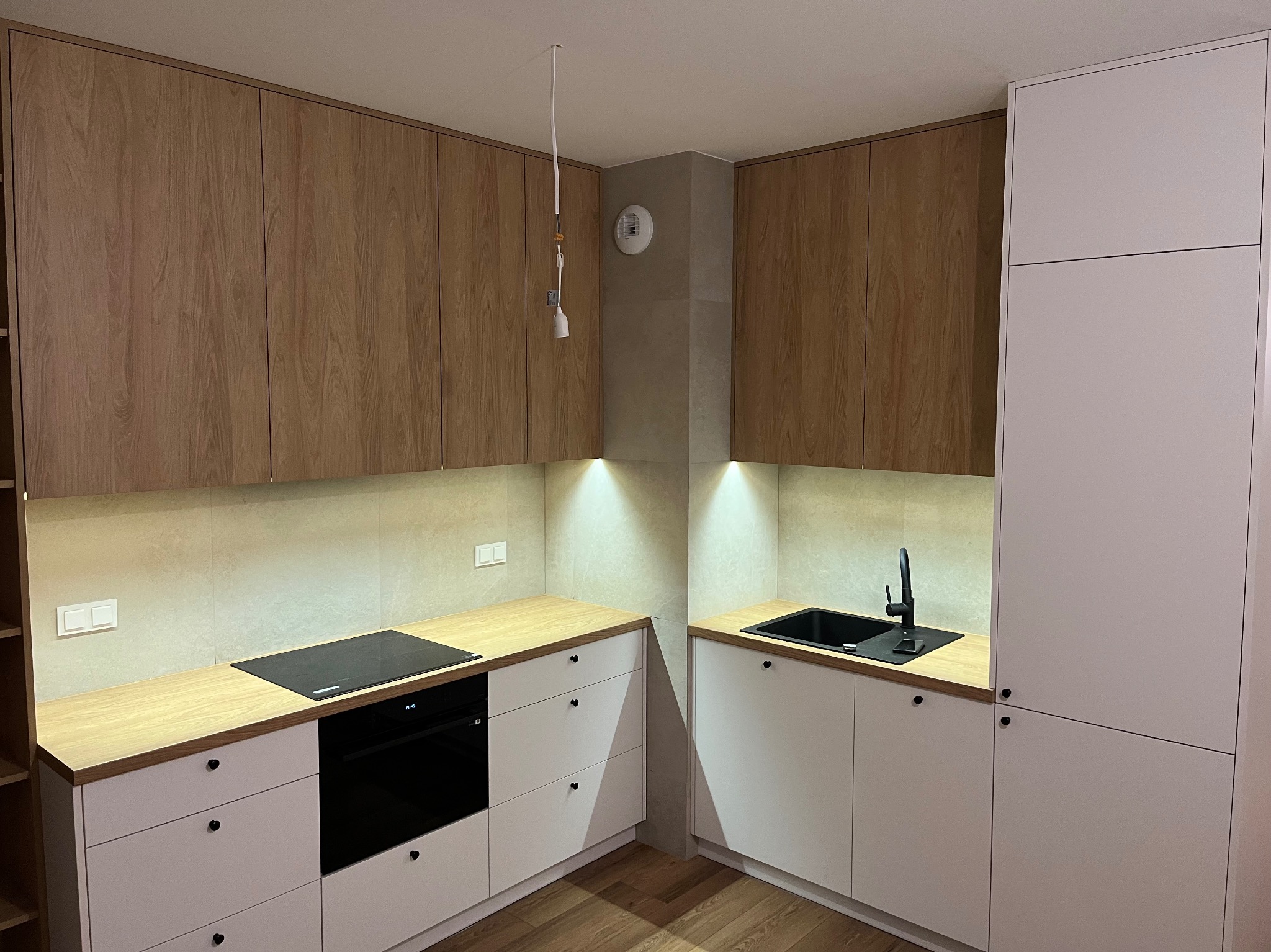 Nowoczesna kuchnia z białymi szafkami dolnymi i drewnianymi górnymi. Blat drewniany, czarny zlew i płyta indukcyjna. Oświetlenie punktowe pod szafkami.