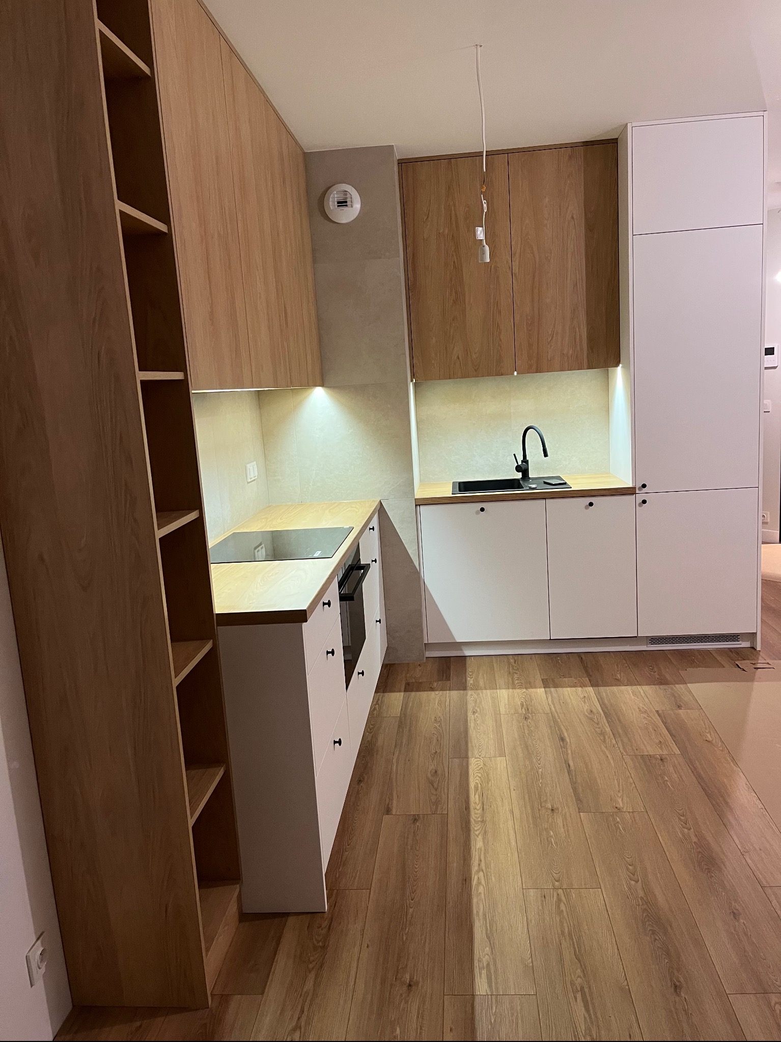 Jasna kuchnia z drewnianymi i białymi frontami, czarną armaturą i płytą indukcyjną. Oświetlenie podszafkowe i drewniana podłoga tworzą przytulny klimat.