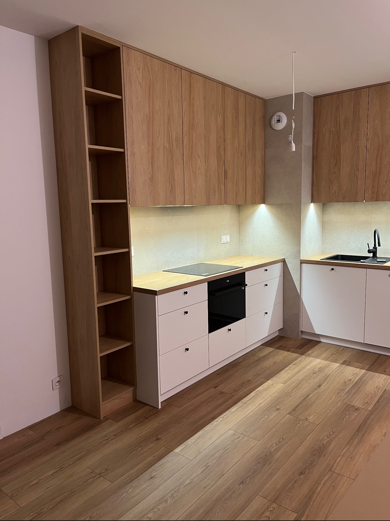 Jasna kuchnia na wymiar z drewnianymi frontami szafek wiszących i białymi dolnymi, z czarnym zlewem i płytą indukcyjną. Dodatkowo wysoki regał z półkami.