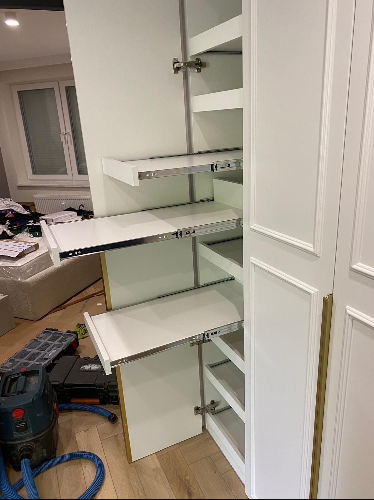 Biała szafa wnękowa z wysuwanymi półkami na metalowych prowadnicach, widoczna z boku, z otwartymi drzwiami. Na podłodze narzędzia i odkurzacz.