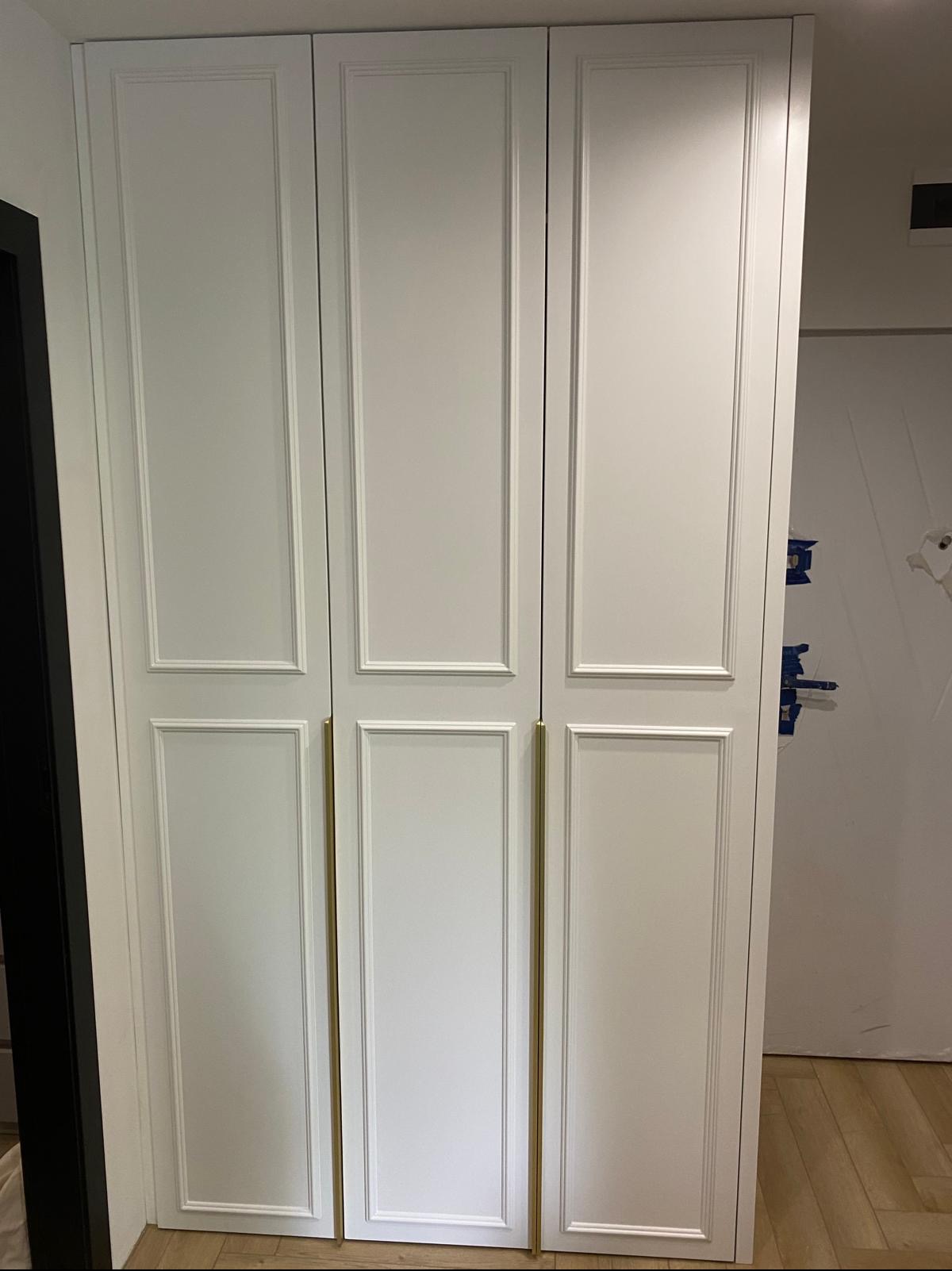 Biała, trzydrzwiowa szafa wnękowa z frezowanymi frontami i długimi, złotymi uchwytami. Widoczny fragment podłogi z jasnego drewna i fragment ściany z drzwiami.