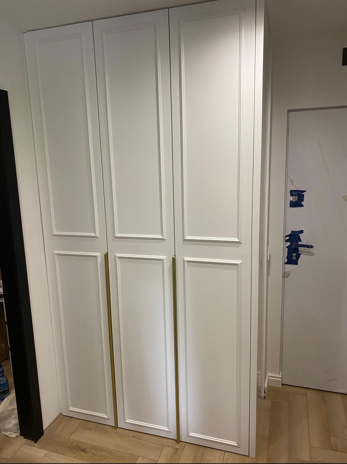 Biała, trzydrzwiowa szafa wnękowa z frezowanymi frontami i złotymi uchwytami, na tle parkietu i fragmentu białej ściany z niebieską taśmą malarską.