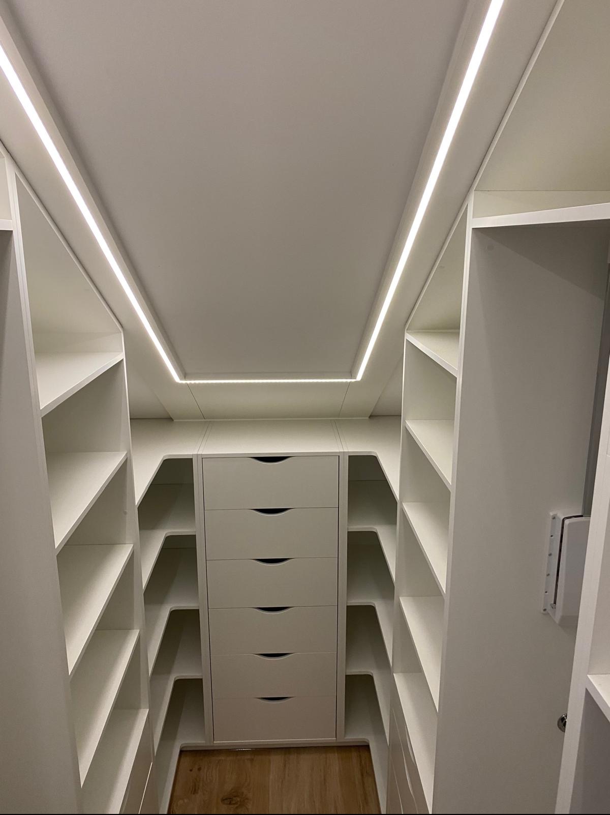Biała zabudowa garderoby pod skosem z oświetleniem LED. Widoczne półki i szuflady. Całość wykonana z płyty laminowanej w kolorze biel alpejska.