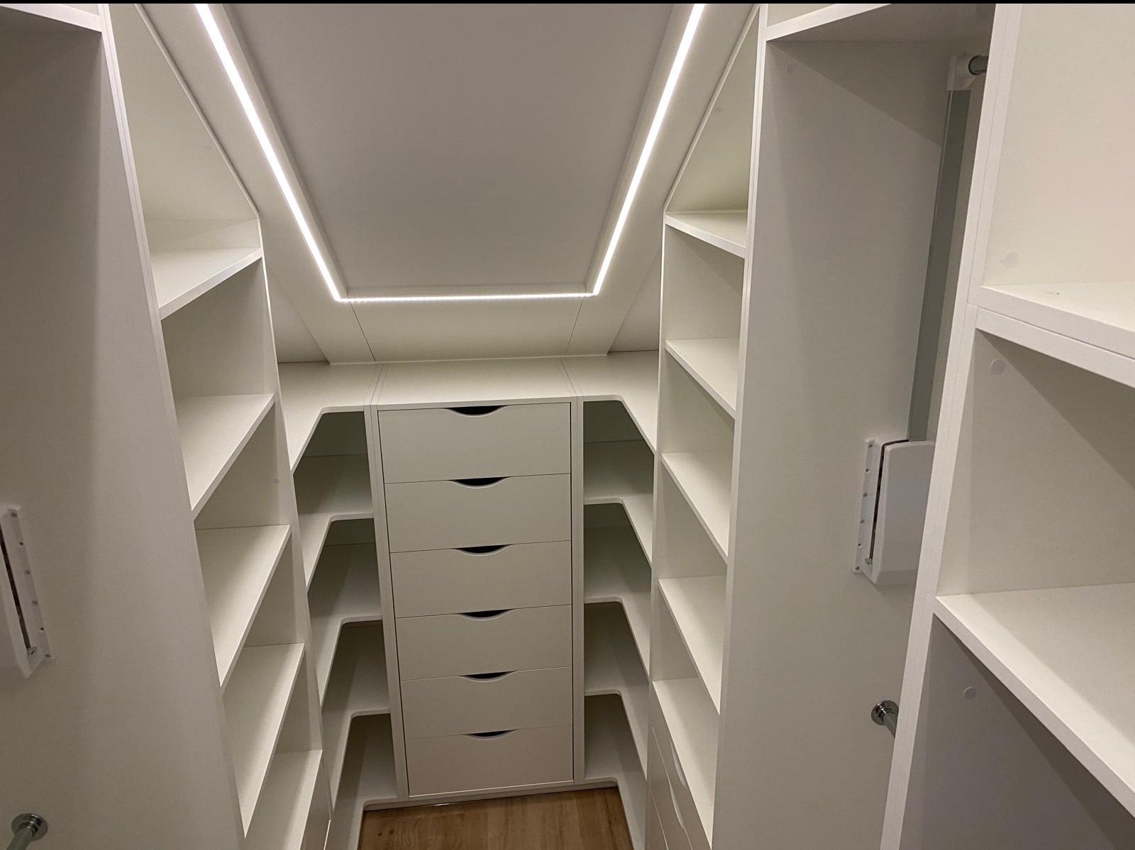 Biała garderoba na wymiar z oświetleniem LED, widok z wnętrza. Szafki z szufladami i półkami, minimalistyczny design, jasne wnętrze, drewniana podłoga.