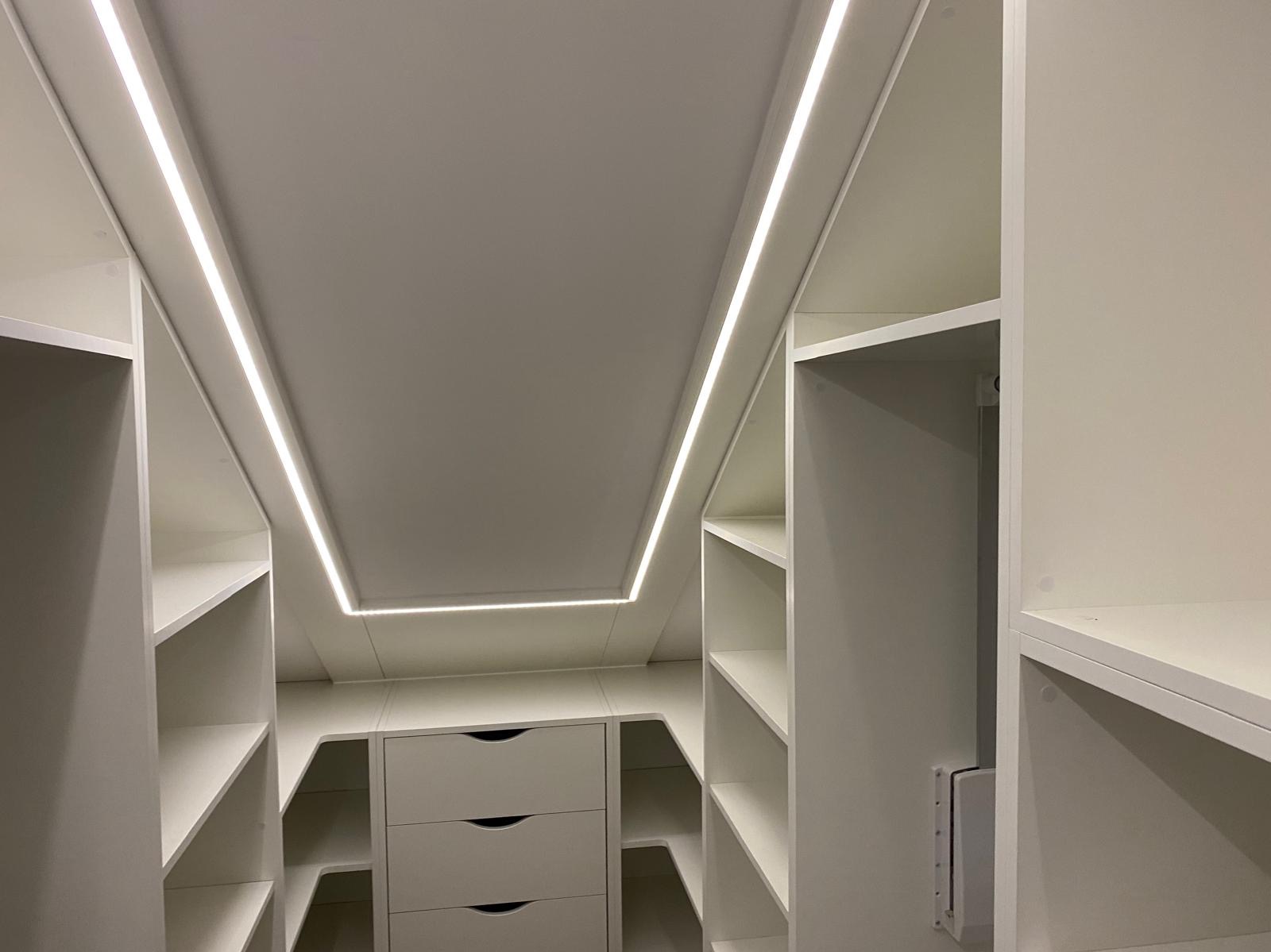 Jasna garderoba na poddaszu z oświetleniem LED. Widok z dołu ukazuje półki i szuflady. Minimalistyczny design, białe wykończenie, nowoczesne oświetlenie.