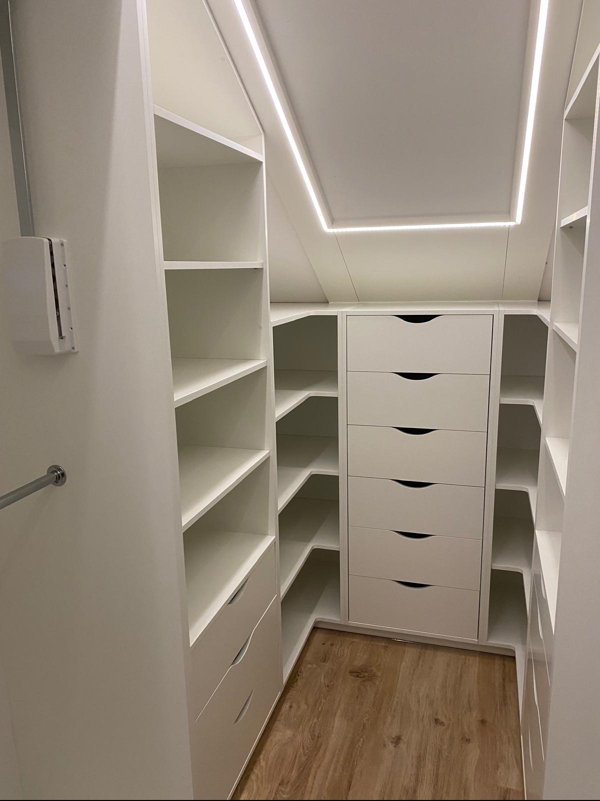 Biała szafa wnękowa z szufladami i półkami, oświetlenie LED w suficie, drewniana podłoga. Realizacja pod skosem, nowoczesny design.