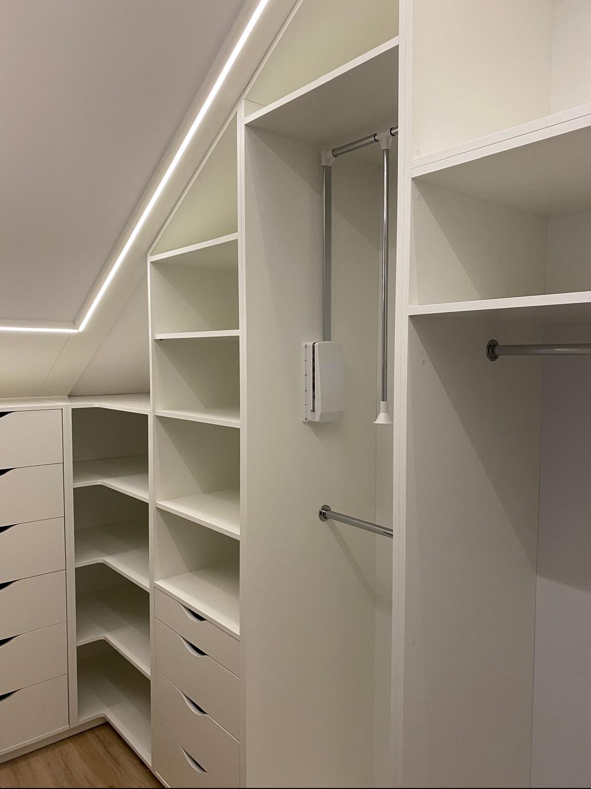 Biała szafa wnękowa z szufladami i półkami, oświetlenie LED w skosie sufitu, drążki na ubrania. Funkcjonalne rozwiązanie do przechowywania w przestrzeni ze skosami.
