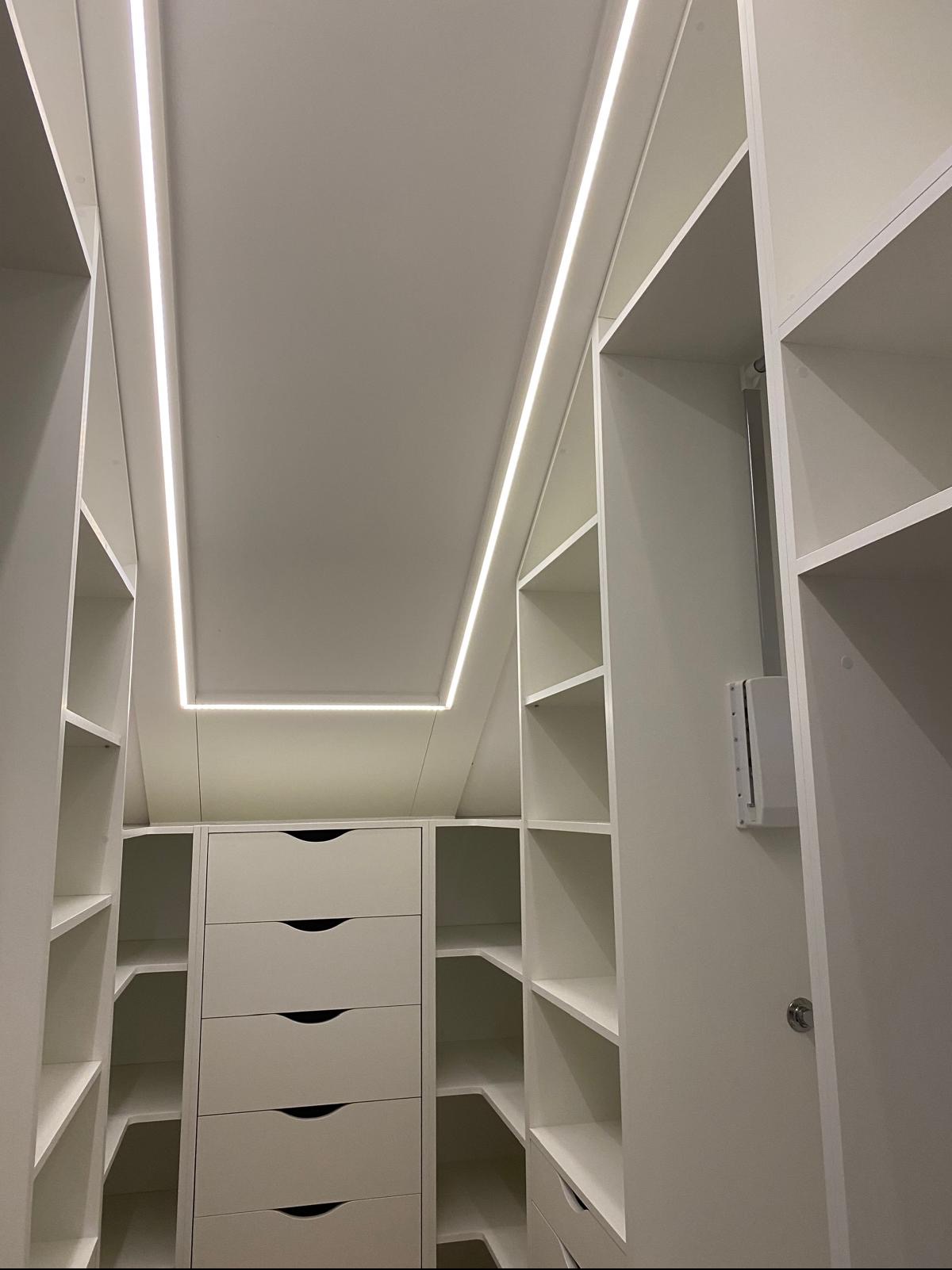 Biała szafa wnękowa z szufladami i półkami, oświetlona ledowym światłem, dopasowana do skosu dachu. Widok z dołu, podkreślający przestrzeń i funkcjonalność.