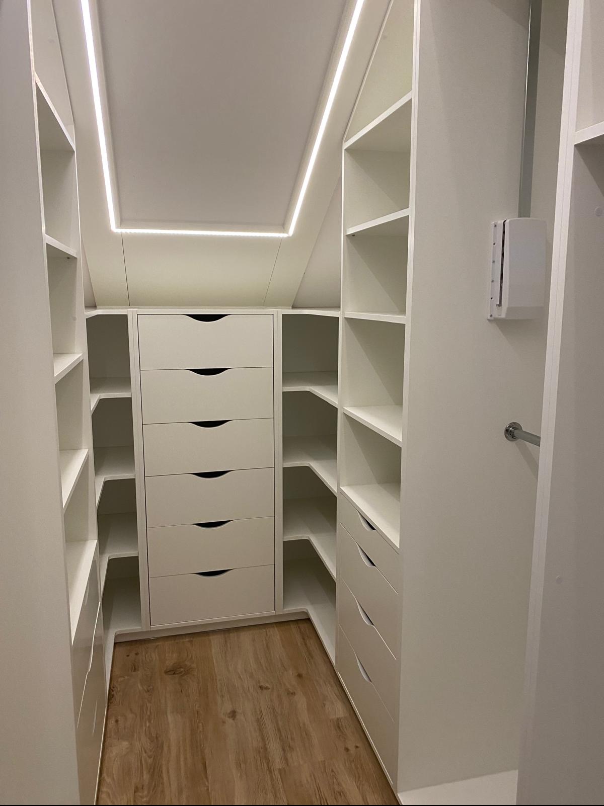 Biała zabudowa wnęki ze szufladami i półkami, oświetlenie LED w suficie, drewniana podłoga. Funkcjonalne rozwiązanie do przechowywania w przestrzeni ze skosami.