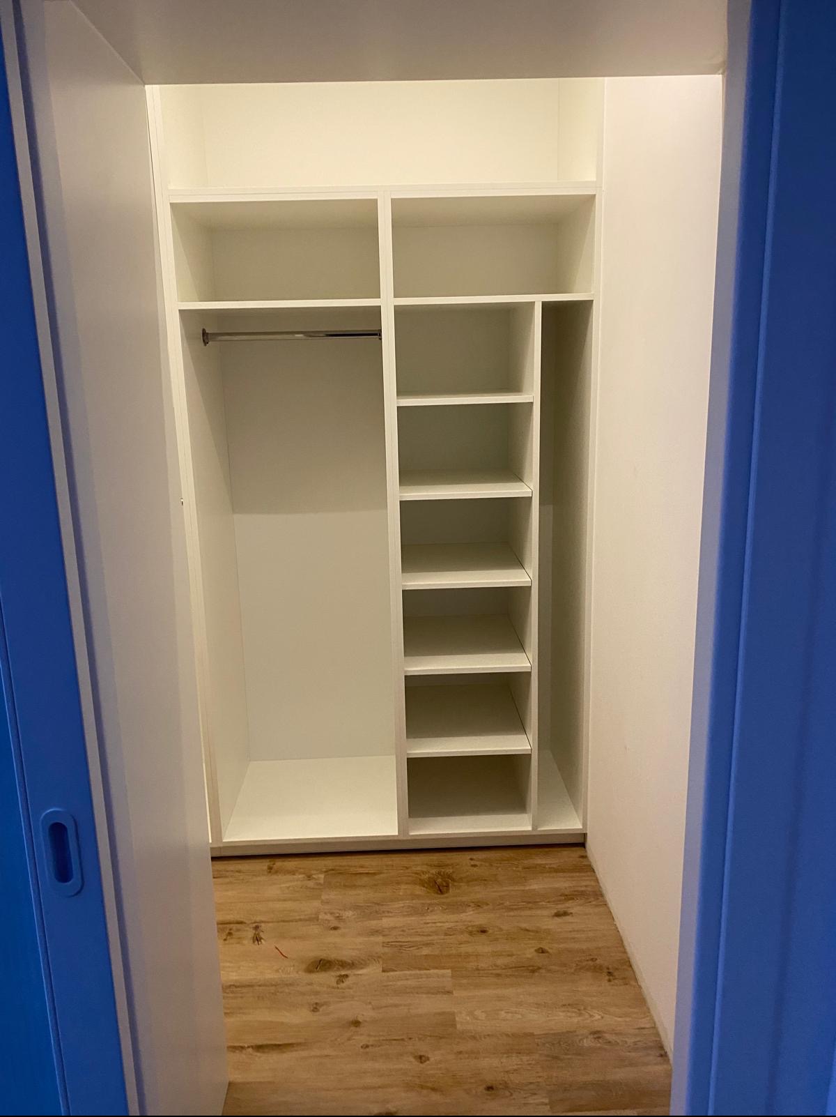 Wnętrze białej szafy wnękowej z drążkiem i półkami, widok z perspektywy wejścia, na podłodze panele imitujące drewno, niebieska ściana po prawej.
