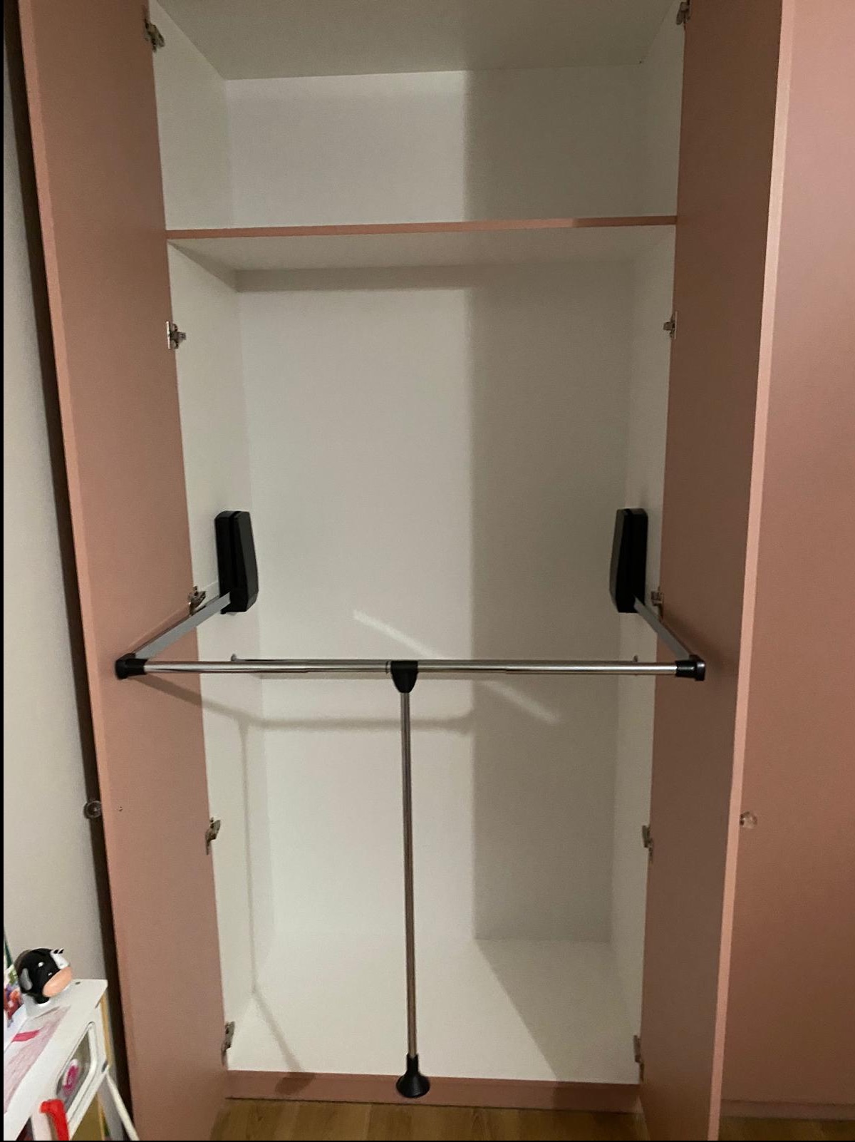 Wnętrze szafy wnękowej z drążkiem na ubrania i półką, fronty w odcieniu pudrowego różu, korpus biały. Widoczne okucia i mechanizm drążka. Ujęcie z dołu, otwarte drzwi.