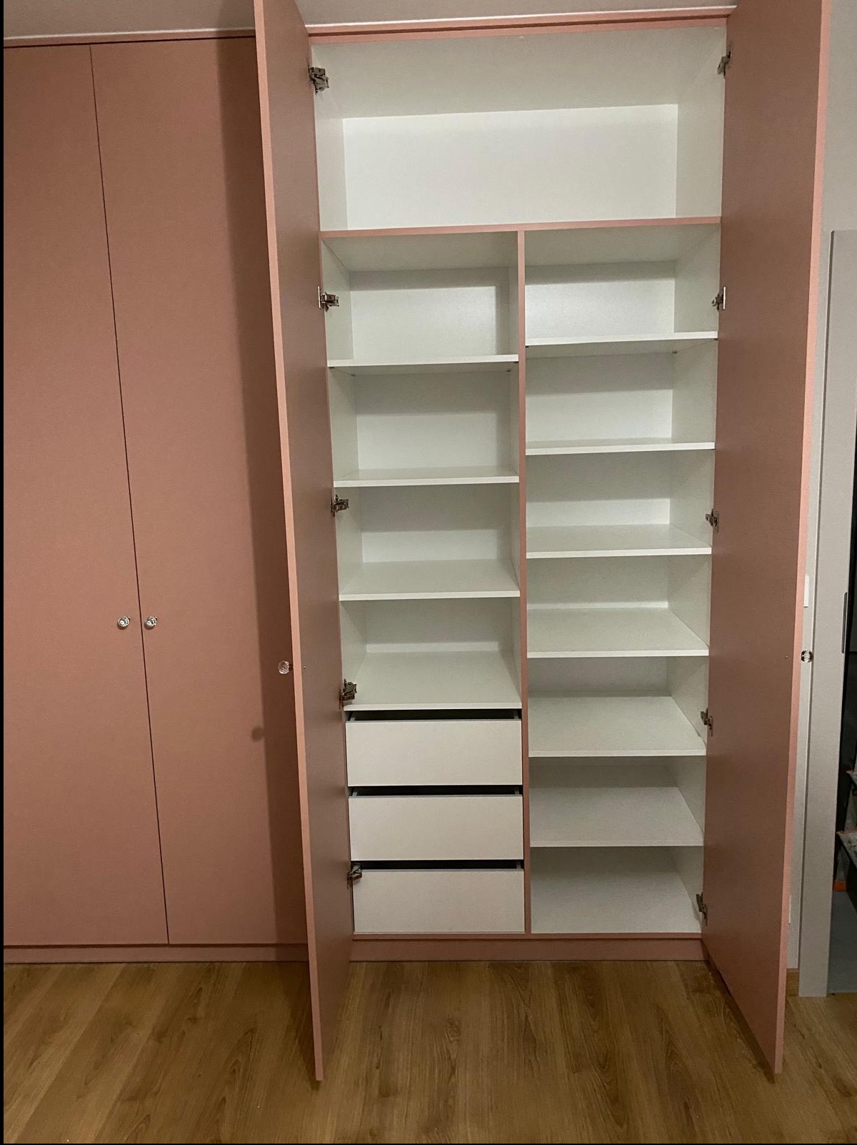 Różowa szafa z otwartymi drzwiami, ukazująca białe półki i szuflady. Widoczne okucia i drewniana podłoga. Minimalistyczny design, jasne wnętrze szafy.