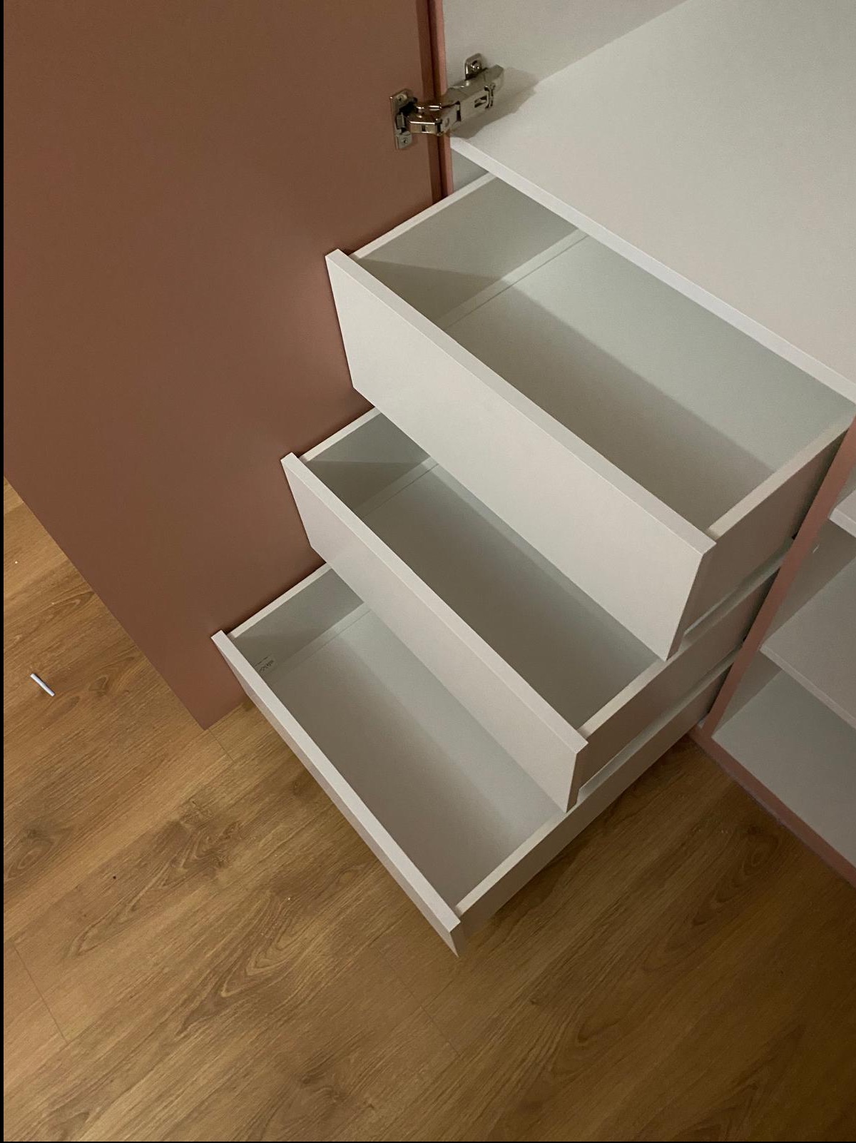 Wnętrze szafy z trzema wysuniętymi, białymi szufladami na tle brązowej ściany i drewnianej podłogi. Widoczne zawiasy i półki wewnątrz szafy.