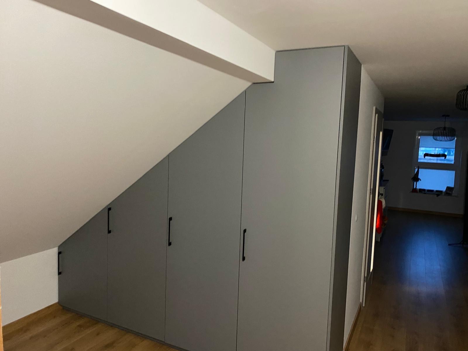 Szara szafa wnękowa z czarnymi uchwytami, dopasowana do skosu poddasza, widoczna z perspektywy korytarza z drewnianą podłogą i oknem w tle.