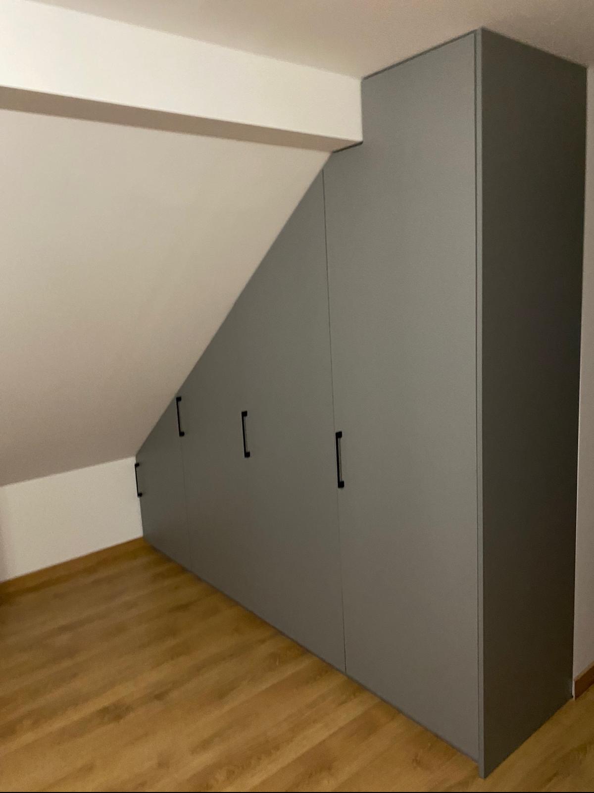 Szara szafa wnękowa z czarnymi uchwytami, dopasowana do skosu poddasza, na drewnianej podłodze. Minimalistyczny design, proste linie, nowoczesne wykończenie.