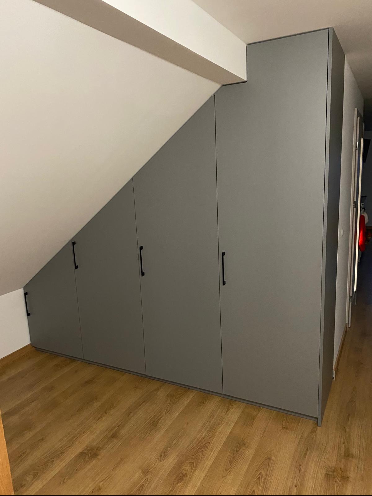 Szara zabudowa szafy wnękowej na poddaszu ze skośnym sufitem, czarne, minimalistyczne uchwyty, drewniana podłoga. Nowoczesny design, funkcjonalne rozwiązanie.