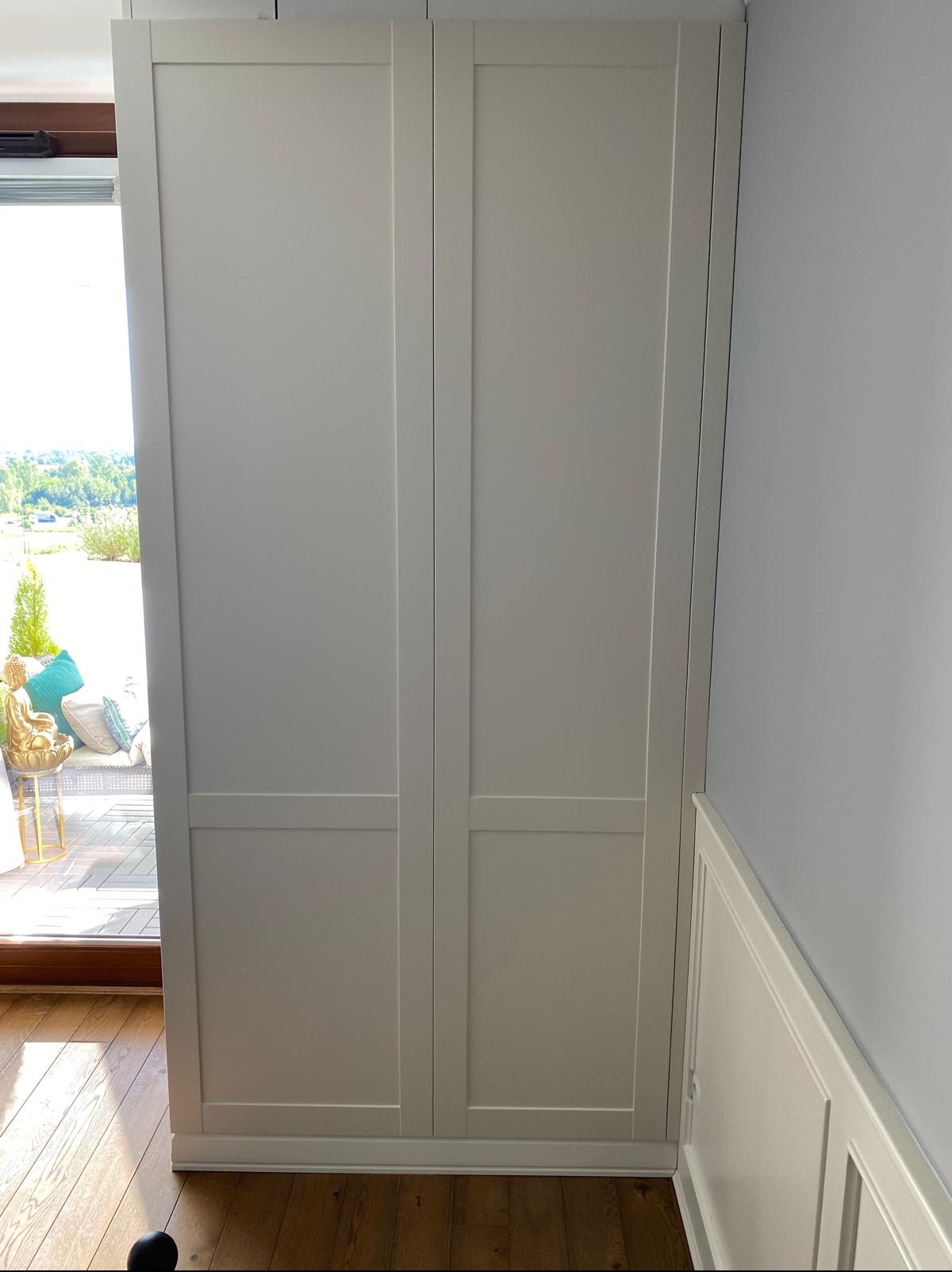 Biała, dwudrzwiowa szafa wnękowa z widokiem na taras z zielenią i dekoracyjnym Buddą. Minimalistyczny design, jasne wnętrze, drewniana podłoga.