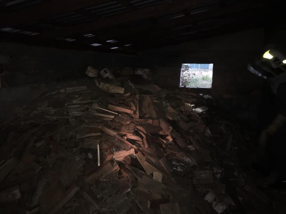 Stos pociętego drewna w ciemnym pomieszczeniu, z widocznym oknem w tle, oświetlenie słabe, elementy osoby w prawym górnym rogu.