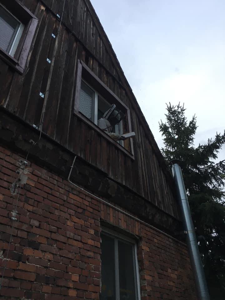 Widok z dołu na drewniany dom z ceglaną dobudówką, z okna wystawiony opryskiwacz ciśnieniowy, prawdopodobnie w trakcie pracy.