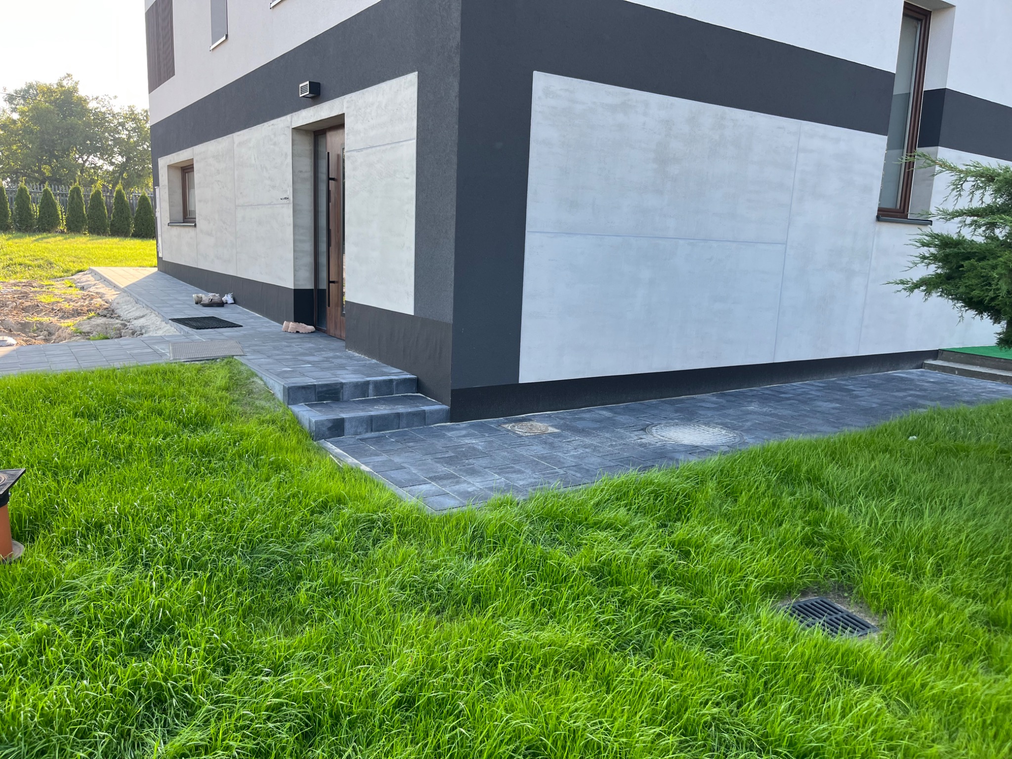 Nowoczesny dom z grafitowo-białą elewacją i ścieżką z szarej kostki prowadzącą do drzwi wejściowych, otoczony soczystą zielenią trawnika.