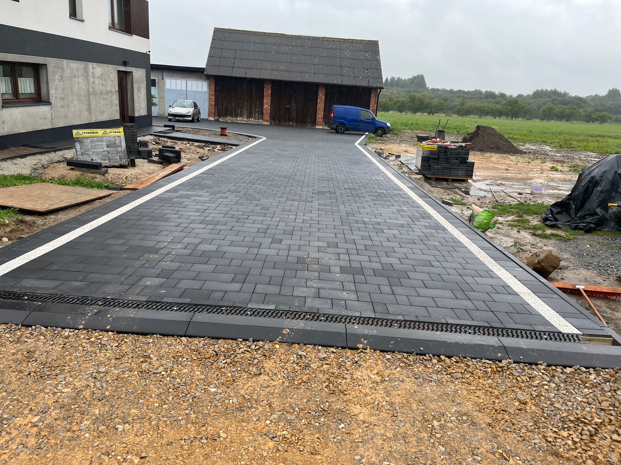 Ukończony podjazd z grafitowej kostki brukowej z białymi pasami, obrzeża z odpływem liniowym, widok na budynki i teren zielony w tle.